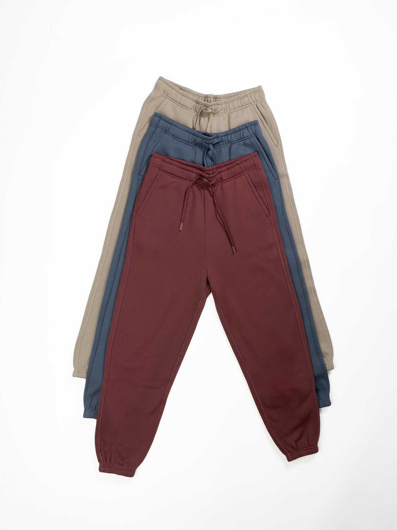Three pairs of Cozy Earth's Men's CityScape Sweatpants are arranged in a fan layout on a white background. The front pair is maroon, the middle pair is blue, and the back pair is beige. All feature drawstrings and elastic cuffs. |Color:Ebony