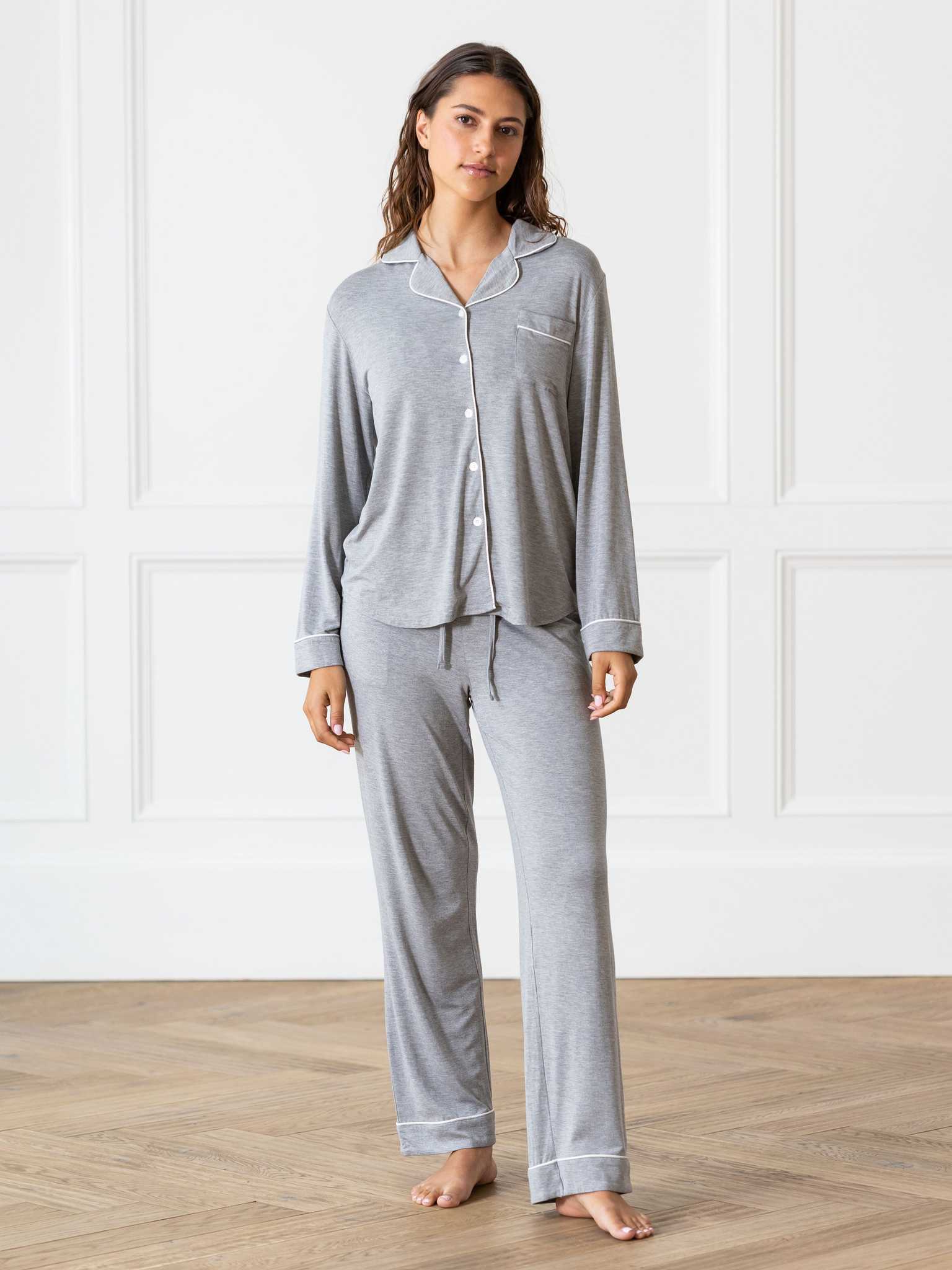A woman is standing on a wooden floor in a room with white paneled walls, dressed in the Women's Bedtime Bundle by Cozy Earth. The gray pajama set features white trim, with the top boasting buttons and a front pocket, while the pants include a drawstring waist. Her long hair flows freely as she stands barefoot. |Color:Grey