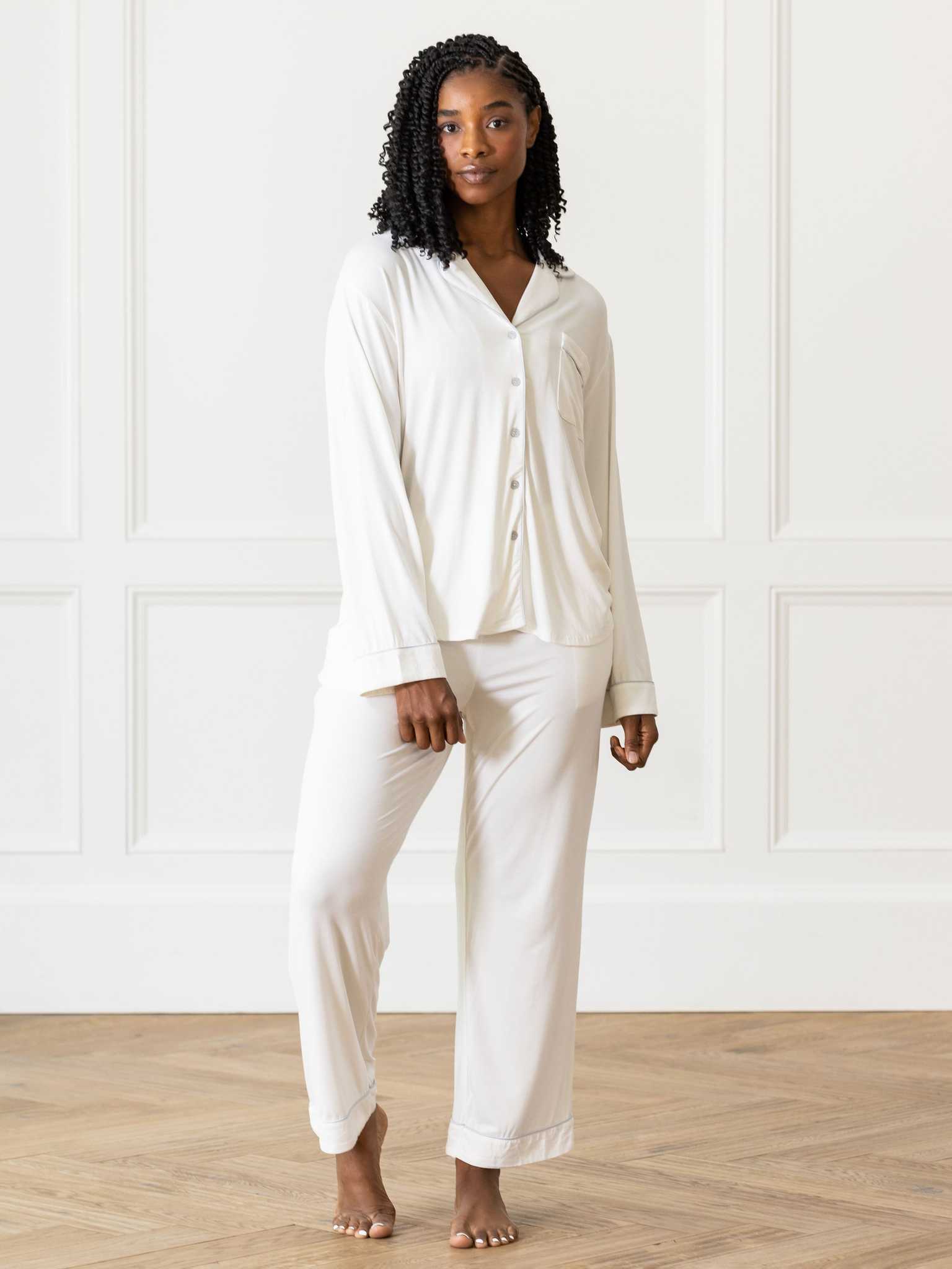 A person with braided hair is standing on a wooden floor against a white paneled wall, wearing the Cozy Earth Women's Bedtime Bundle, which consists of a cream-colored long-sleeved button-up top and matching pants. |Color:Ivory