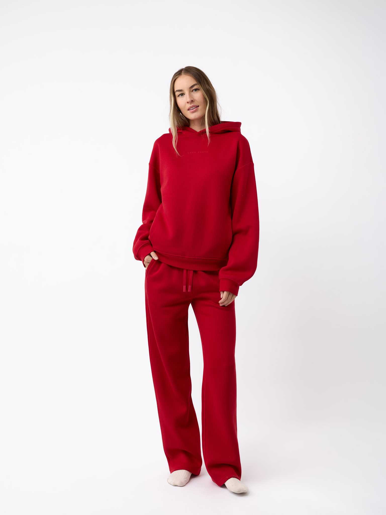 A person stands against a plain white background wearing a matching Women's CityScape Hoodie by Cozy Earth and sweatpants. They have long hair and are looking towards the camera with a relaxed posture. |Color:Crimson