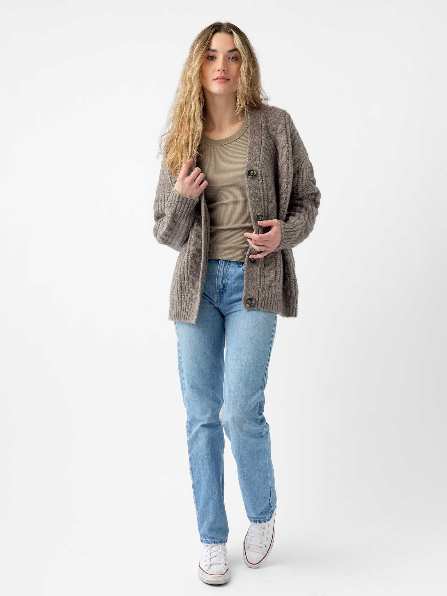 A person with long, wavy hair wears a beige top, light blue jeans, and white sneakers. They stand against a plain white background, gazing directly at the camera with one hand on their Cozy Earth Oversized Cable Knit Cardigan. |Color:Greige
