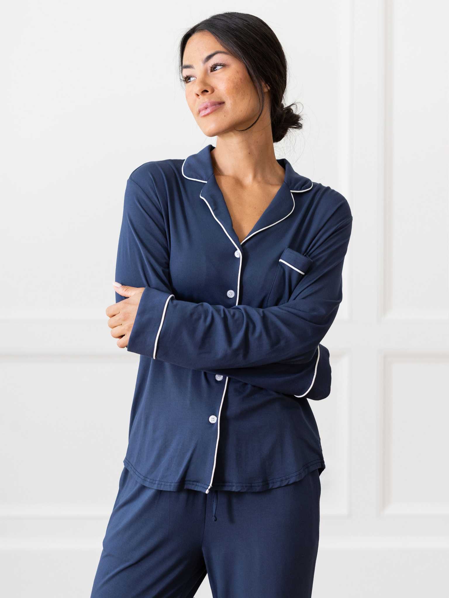 A person with long dark hair tied back is wearing the Women’s Bedtime Bundle from Cozy Earth, a dark blue pajama set with white piping. The top features a collar, buttons, and a chest pocket. The background features white-paneled walls. The person stands with their arms crossed, looking to the side. |Color:Navy