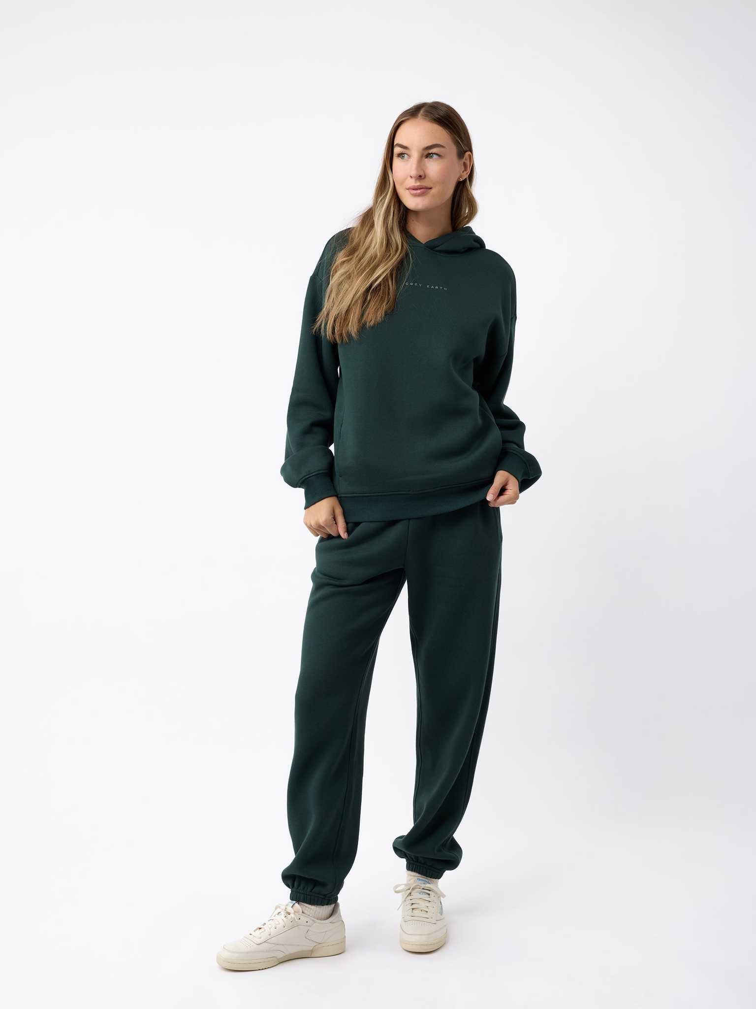 A woman stands confidently against a plain, white background, wearing a dark green CityScape Hoodie by Cozy Earth and matching jogger pants. She has long hair and is looking to the side, with her hands in her pockets and light-colored sneakers on her feet. |Color:Spruce