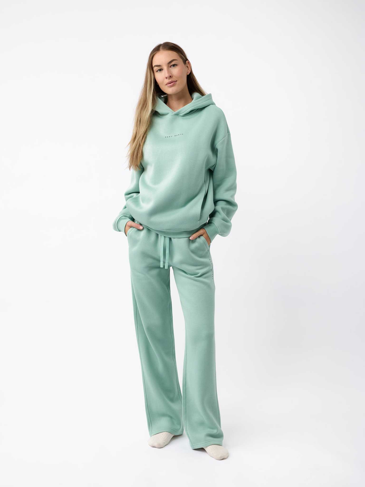 A person dressed in a mint green Women's CityScape Hoodie by Cozy Earth and matching joggers stands with hands in pockets against a plain white background. They have long, light brown hair and are sporting cream-colored shoes. |Color:Wintergreen