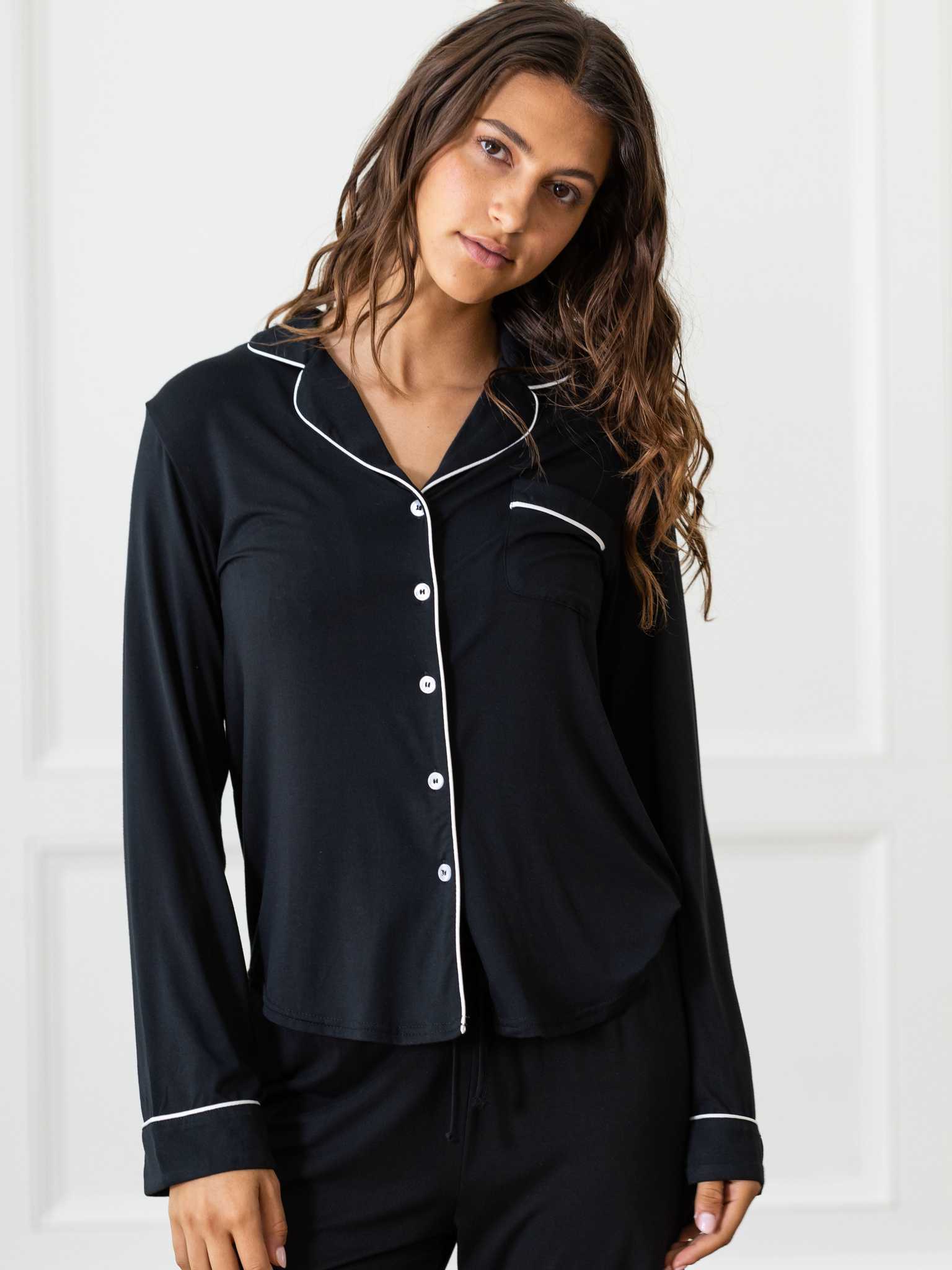 A person with long, wavy hair stands indoors wearing Cozy Earth's Women's Bedtime Bundle, featuring a black pajama top adorned with white piping along the collar, button placket, and cuffs. The top includes a chest pocket and is paired with matching black pajama pants. The background showcases a light-colored wall with decorative trim. |Color:Black