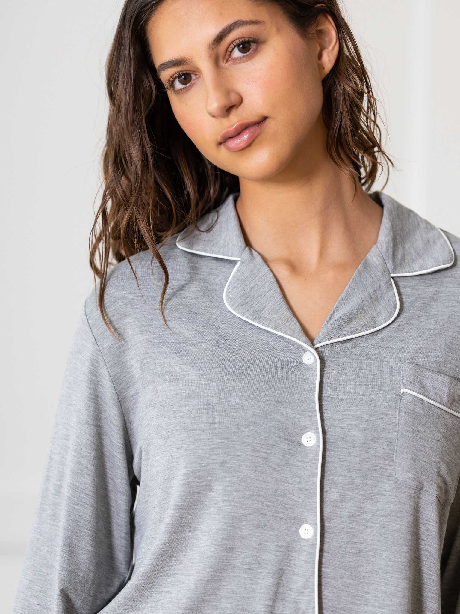 A woman with wavy brown hair is wearing the Women's Bedtime Bundle by Cozy Earth, featuring a light gray pajama top with white piping and buttons. She stands in front of a plain white background, looking at the camera with a neutral expression. |Color:Grey