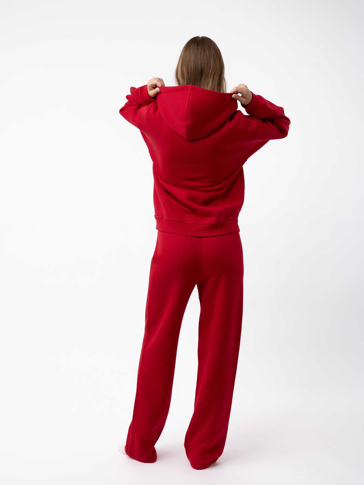 A person stands facing away, wearing a matching red Women's CityScape Hoodie and pants from Cozy Earth. They are holding the hood of the hoodie, set against a plain white background. |Color:Crimson