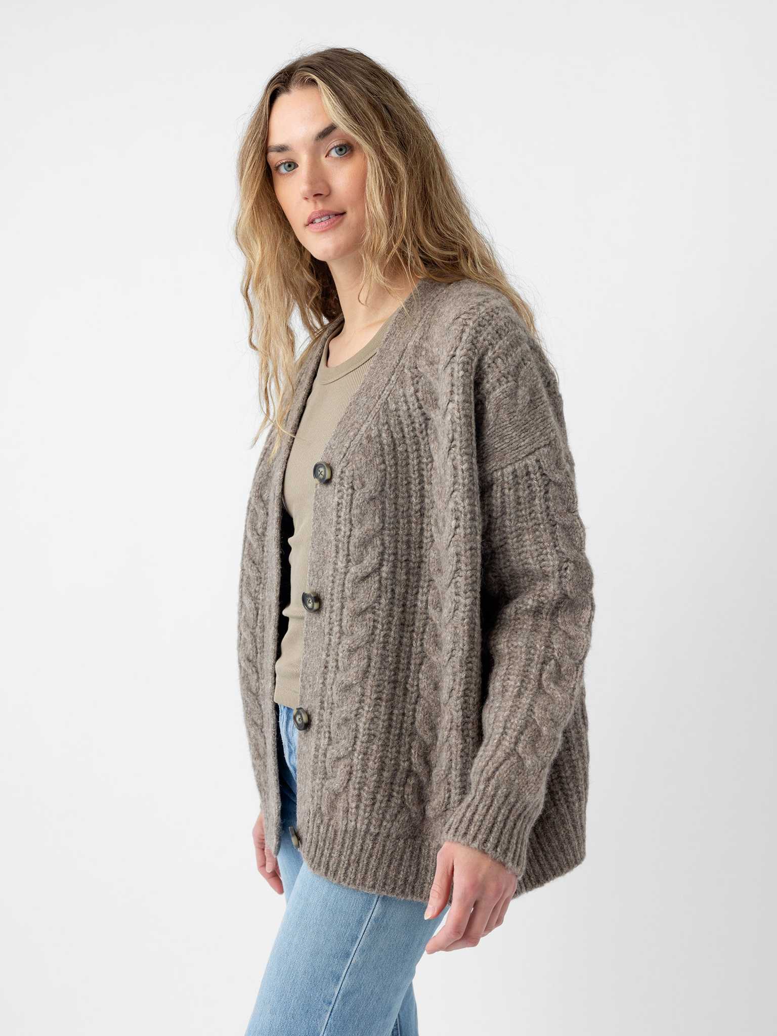 A person with long hair is wearing the "Oversized Cable Knit Cardigan" by Cozy Earth, along with a light shirt. They are standing against a plain background, also dressed in light blue jeans, and gazing toward the camera. |Color:Greige
