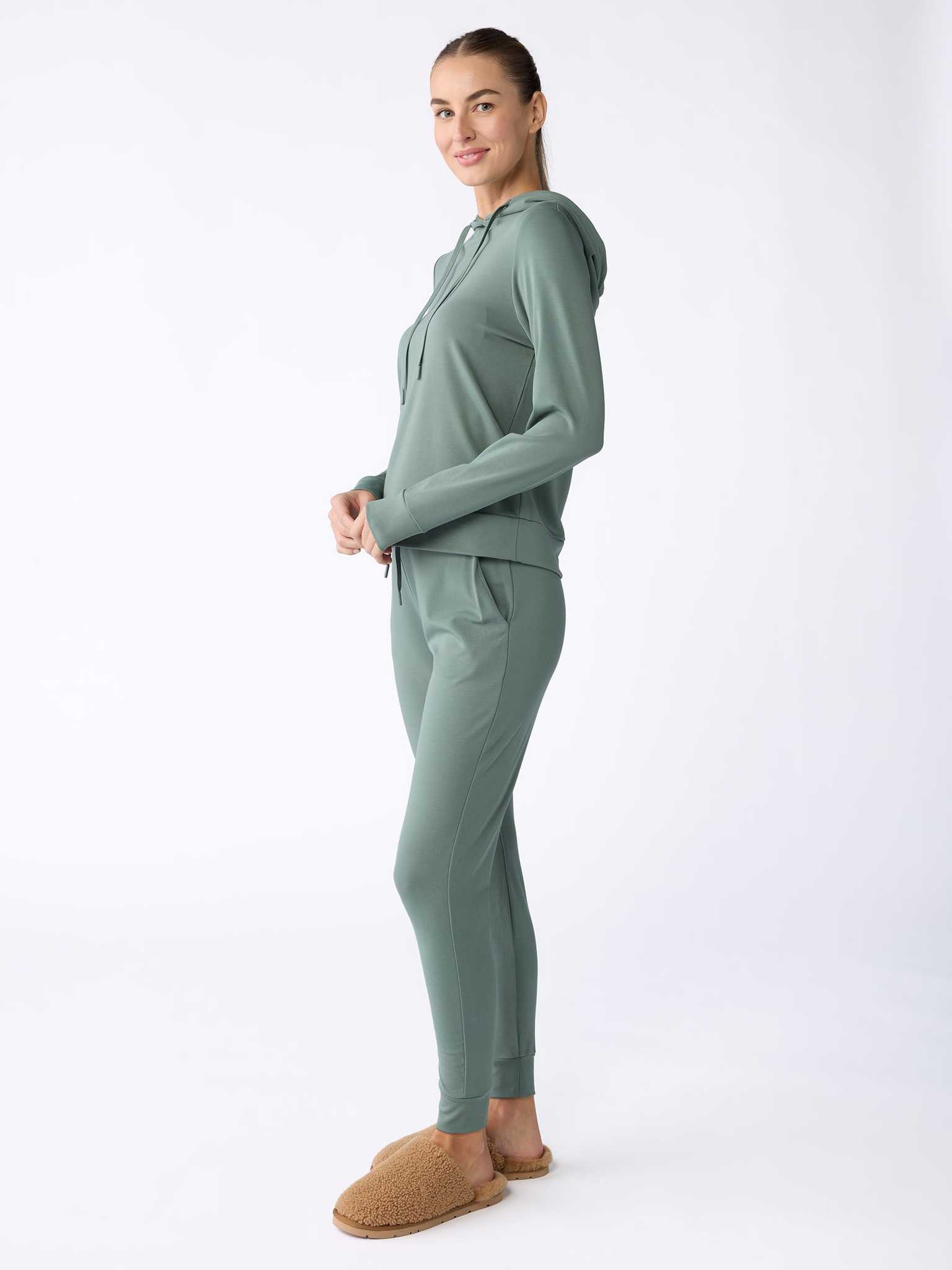 A woman in a Women's Ultra-Soft Bamboo Hoodie by Cozy Earth, paired with jogger pants, stands smiling against a plain white background. Her hair is styled in a ponytail, and she completes her look with fluffy brown slippers. |Color:Oakmoss