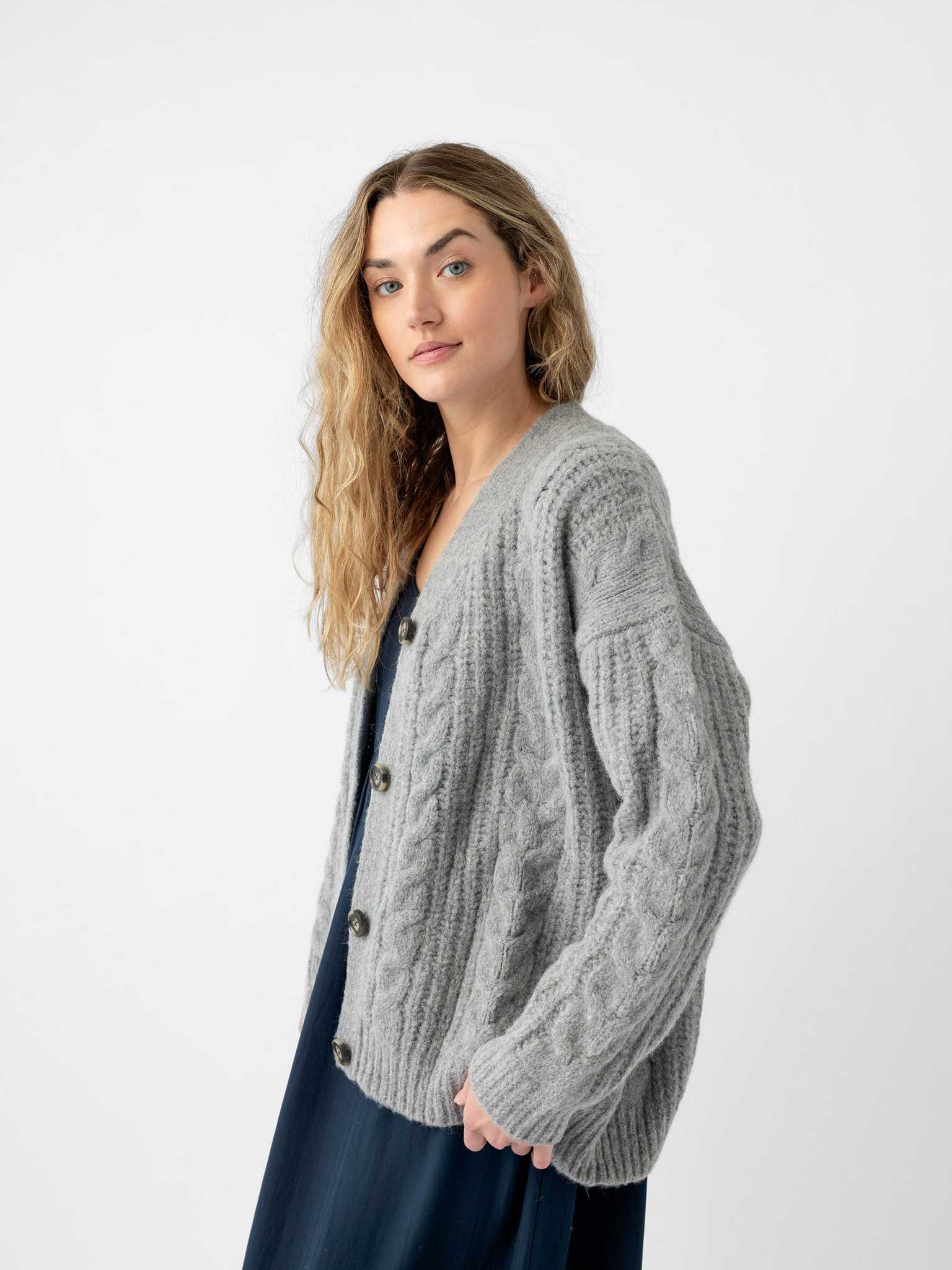 A person with long, curly hair is wearing the Oversized Cable Knit Cardigan by Cozy Earth in gray over a dark dress. They are standing against a plain, white background and looking towards the camera. |Color:Oxford
