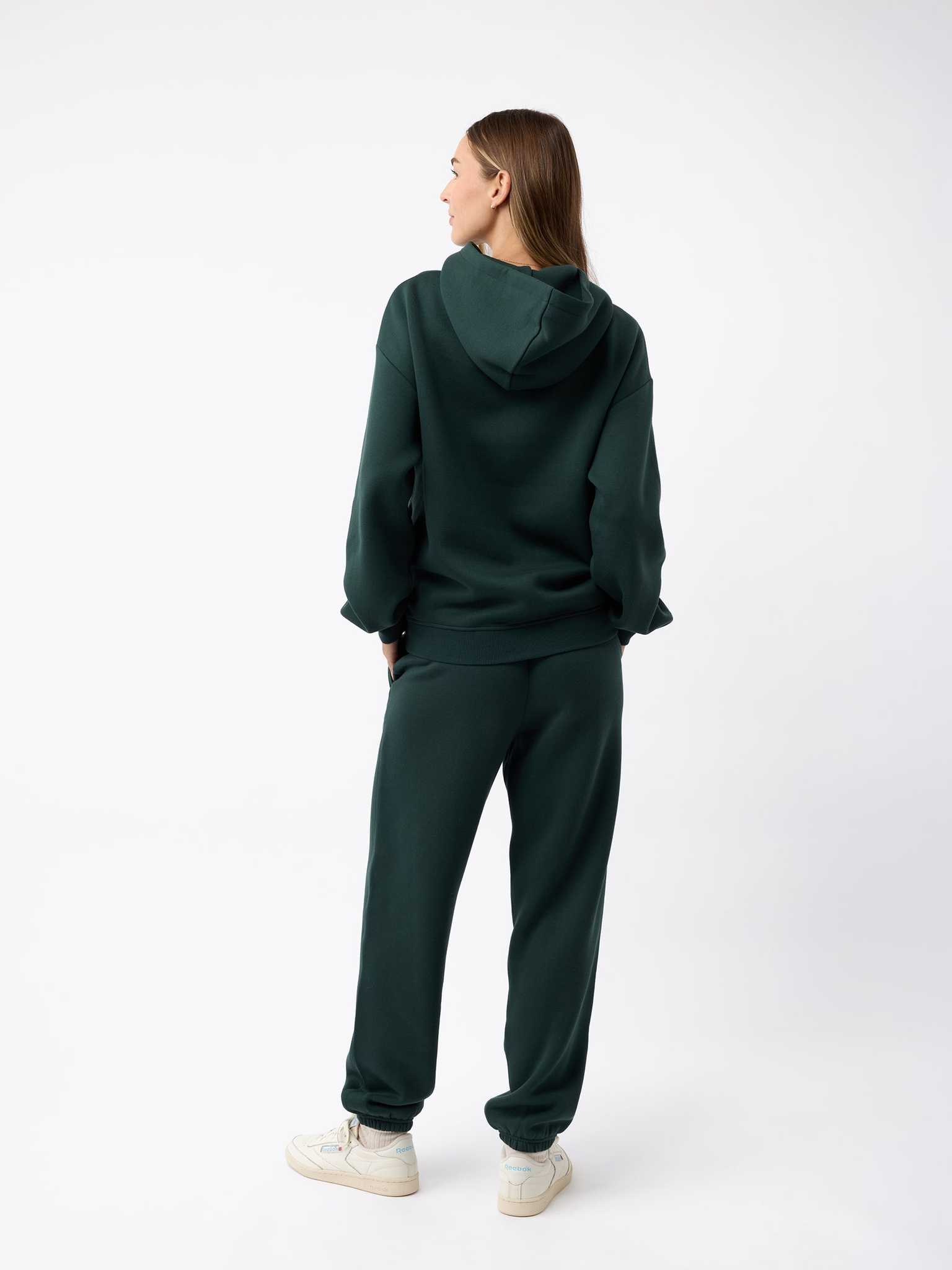 A person stands facing away, wearing a dark green Women's CityScape Hoodie by Cozy Earth with matching sweatpants. They have long hair and are also wearing white sneakers. The background is plain white. |Color:Spruce
