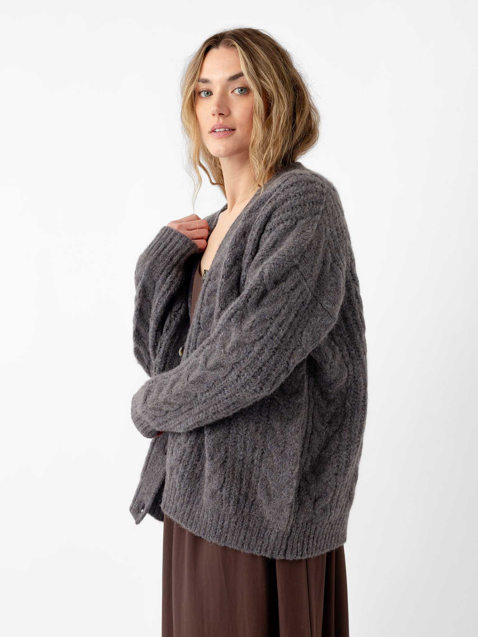 A person with long hair stands against a white background, wearing Cozy Earth's Oversized Cable Knit Cardigan in gray over a brown dress. They maintain a neutral expression as they look towards the camera, gently holding the cardigan collar with one hand. |Color:Thunder