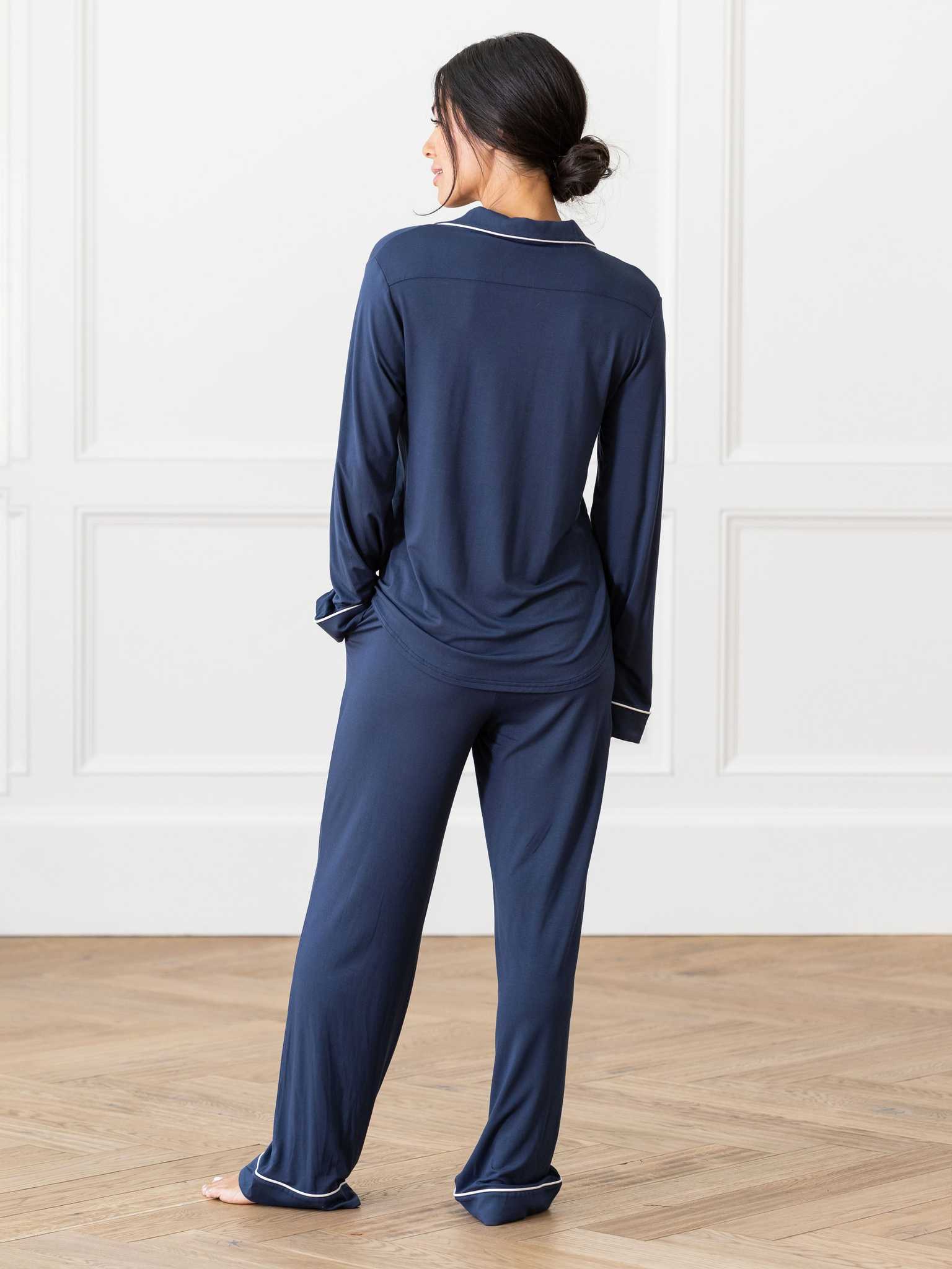A person with dark hair tied in a low bun stands barefoot on a wooden floor facing away from the camera, dressed in Cozy Earth's Women's Bedtime Bundle, a matching dark blue pajama set with long sleeves and pants. The background features white walls with decorative paneling. |Color:Navy