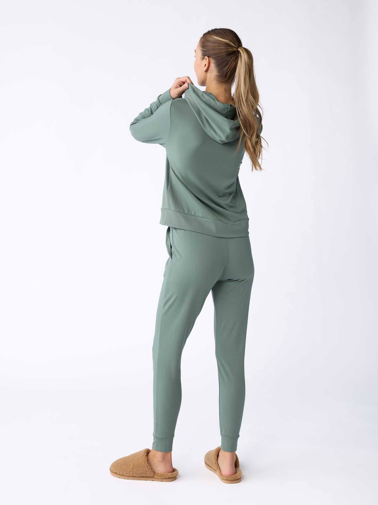 A woman with long hair stands facing away from the camera on a white background, wearing a cozy green Women's Ultra-Soft Bamboo Hoodie from Cozy Earth and tan slippers. |Color:Oakmoss