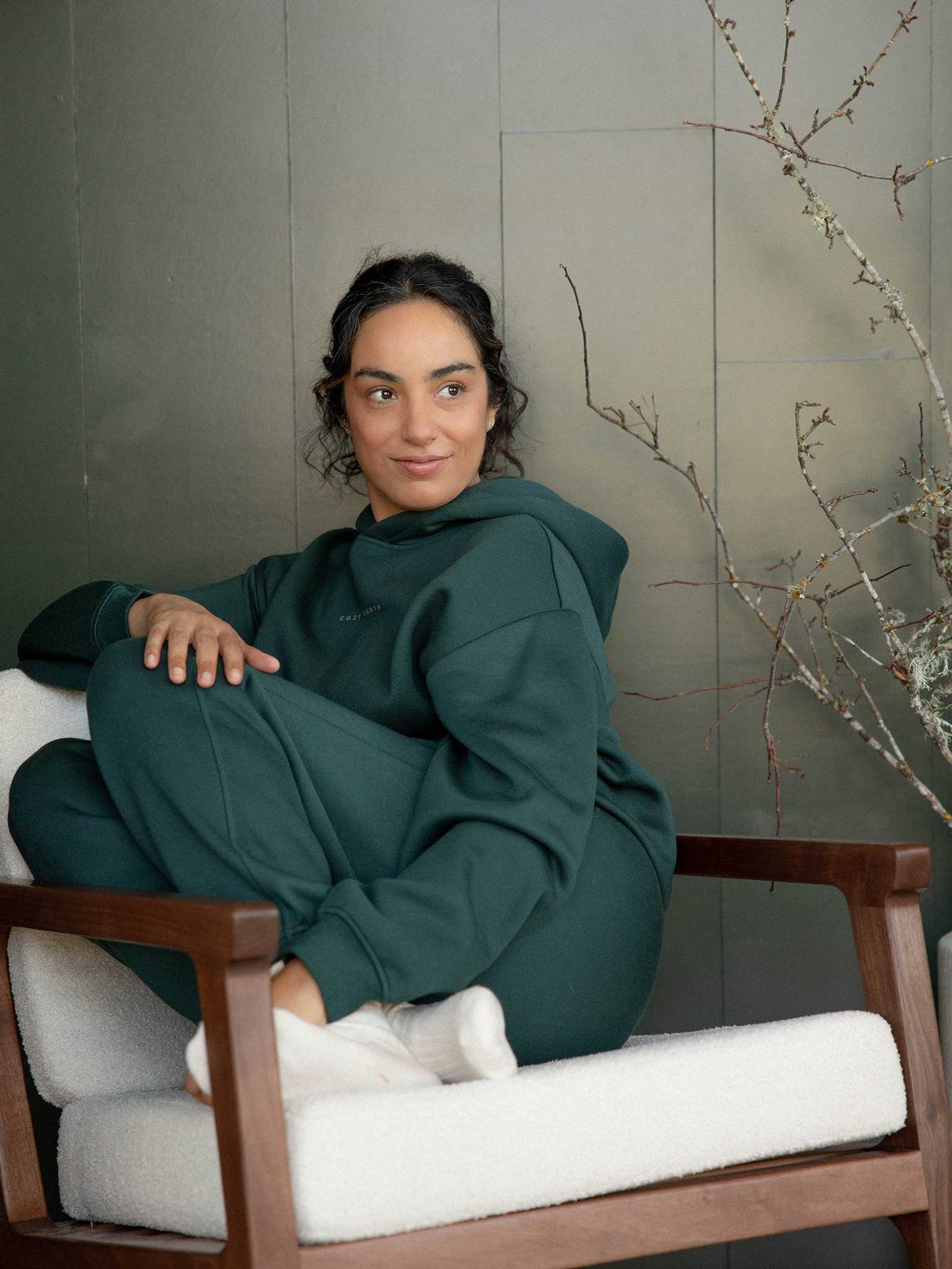 A person with dark hair sits comfortably on a white cushioned chair, wearing the Cozy Earth's Women's CityScape Hoodie paired with sweatpants. They have a relaxed expression as they gaze off-camera. In the background, a bare branch decorates the scene against a greenish wall. |Color:Spruce