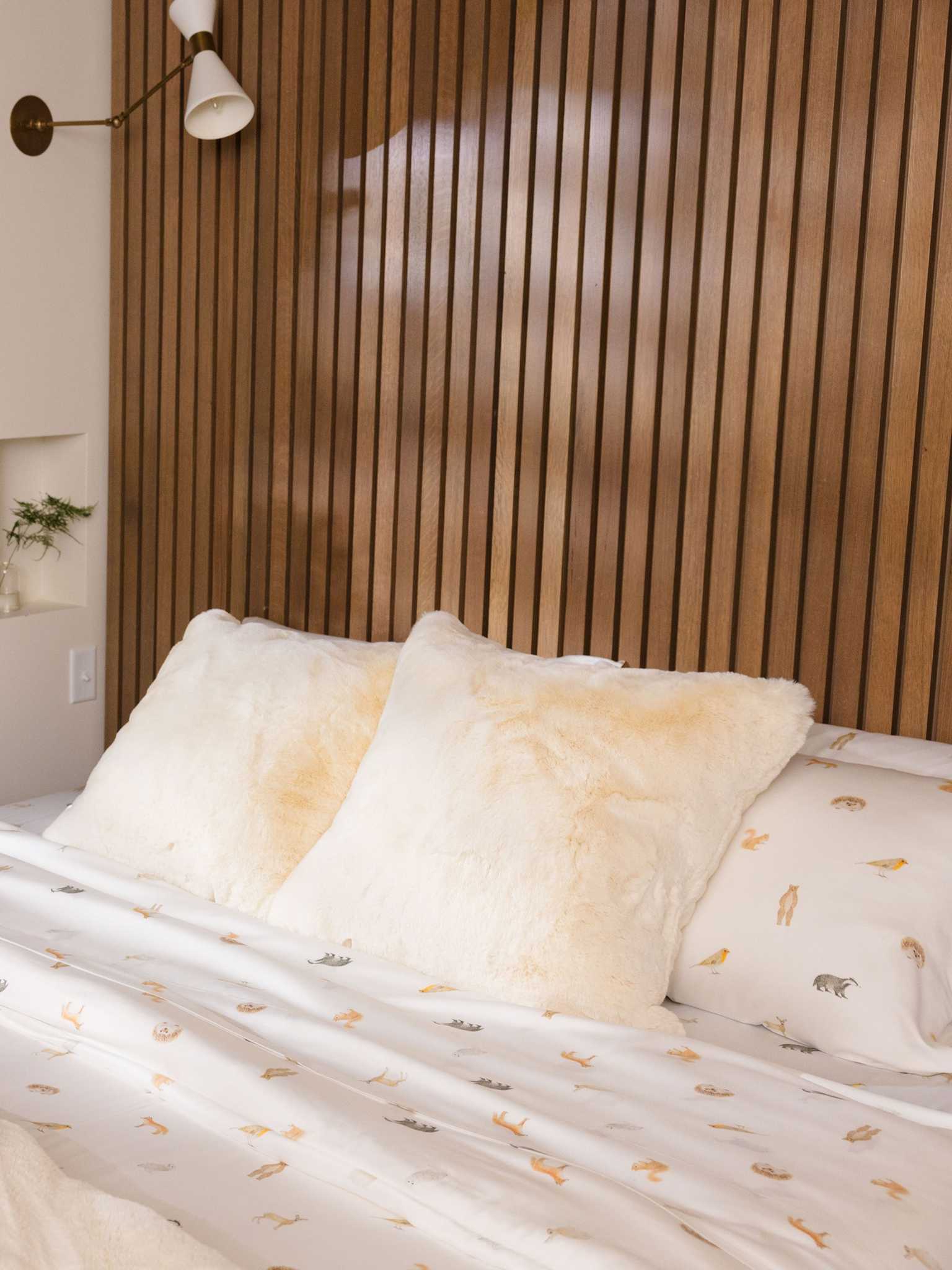 A cozy bed featuring Cozy Earth's Bamboo Duvet Cover, complemented by two large, fluffy cream-colored pillows against a wooden slat headboard. The bedspread boasts a minimalist illustration print, and there's a wall-mounted white lamp on the left side. |Color:Woodland Creatures