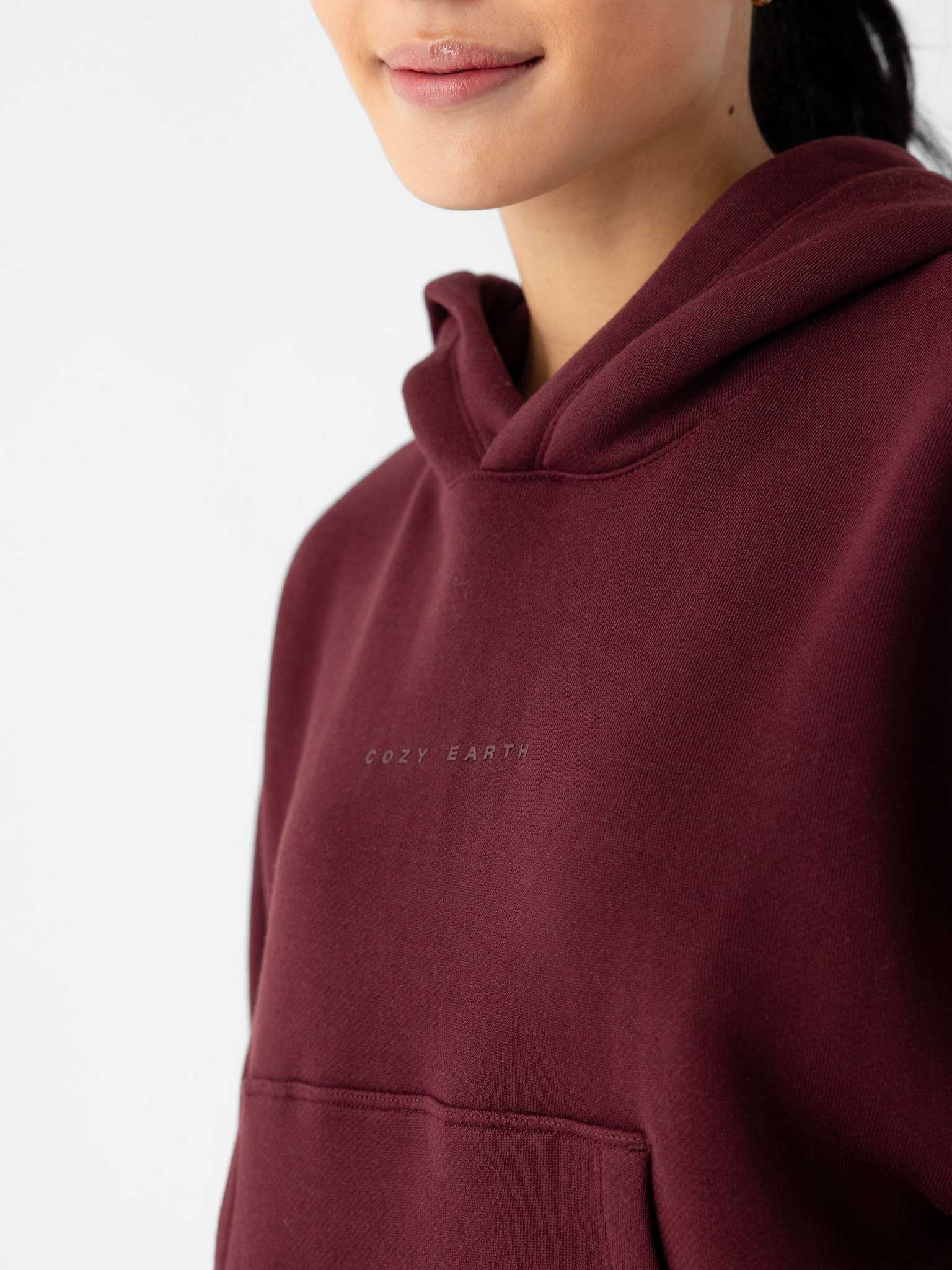 Close-up of a person wearing a Women's CityScape Cropped Hoodie in maroon with the text "Cozy Earth" printed on the chest. Only the lower part of the person's face is visible, along with the upper portion of the hoodie and its front pocket. The background is plain white. |Color:Burgundy