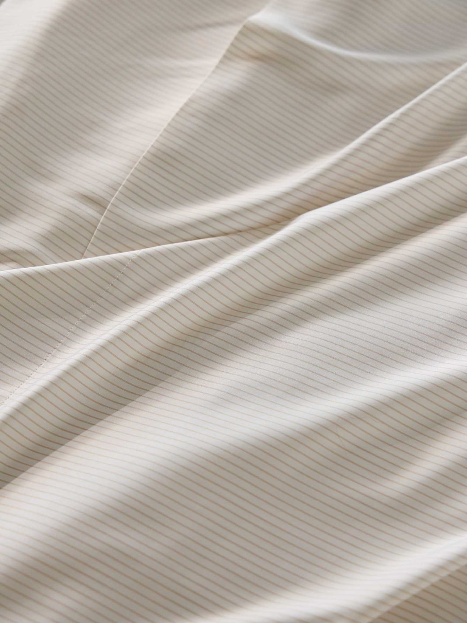Close-up of the Bamboo Duvet Cover by Cozy Earth, showcasing its smooth, white fabric adorned with thin, light beige stripes running diagonally. The duvet cover is gently folded, creating soft waves and subtle shadows. |Color:Driftwood Pinstripe