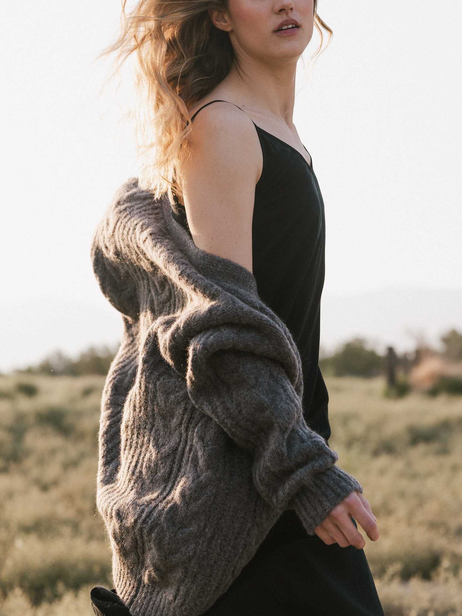 A person with wavy hair walks through a field, wearing a black dress and an Oversized Cable Knit Cardigan by Cozy Earth draped over their shoulder. The scene is serene with a soft, warm light. |Color:Thunder