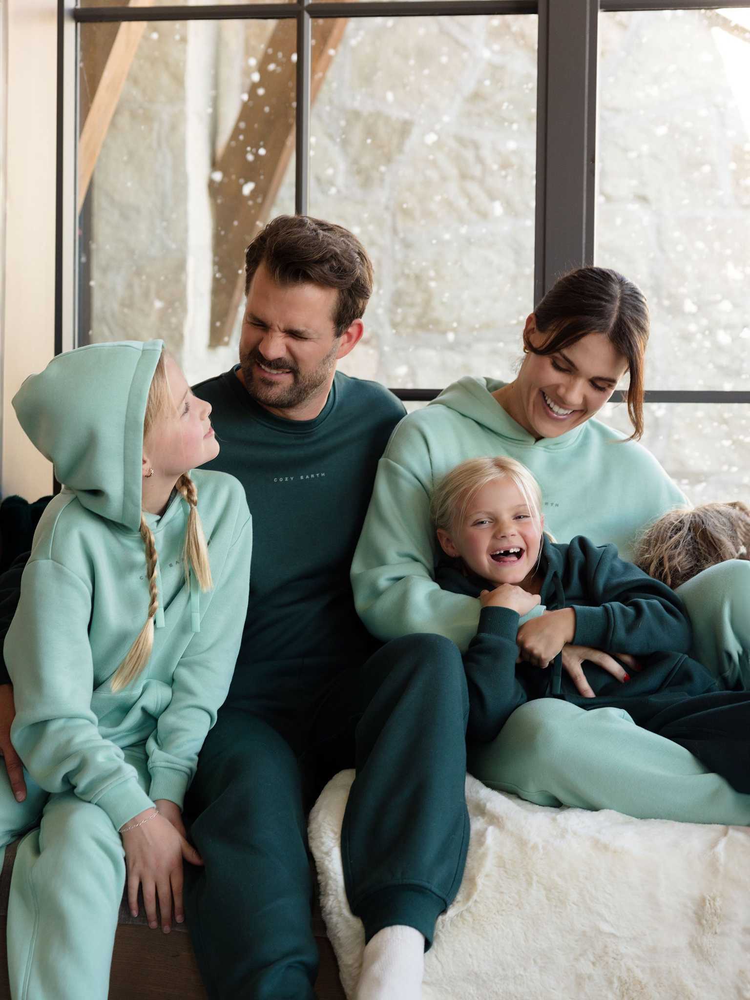 A family of four is gathered indoors, smiling as they wear matching teal and green Women's CityScape Hoodies from Cozy Earth, paired with sweatpants. The cozy and cheerful scene is complete with a snowy landscape visible through the large windows in the background. |Color:Wintergreen