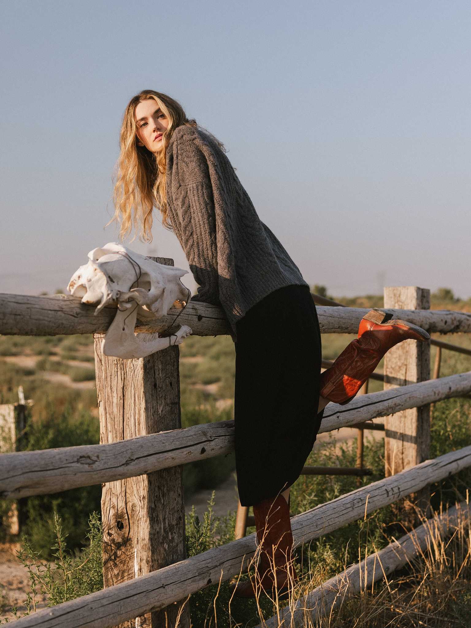 A person with long hair, dressed in an Oversized Cable Knit Cardigan by Cozy Earth, a black skirt, and red boots, leans on a wooden fence while gazing into the distance. A white skull is positioned next to them in this rural setting under a clear blue sky. |Color:Thunder