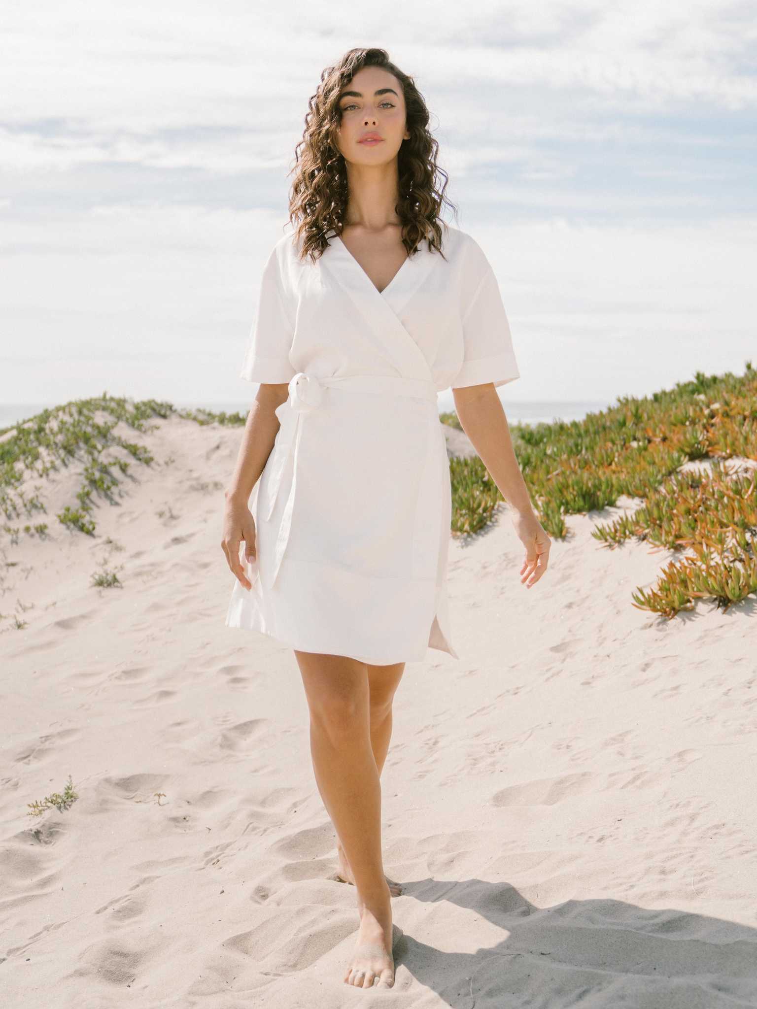 A person with curly hair walks barefoot on a sandy beach path, adorned in the Women's Cozy Earth Coastal Comfort Wrap Dress by Cozy Earth. The sky is partly cloudy, and the path is flanked by lush green shrubs and plants. |Color:White