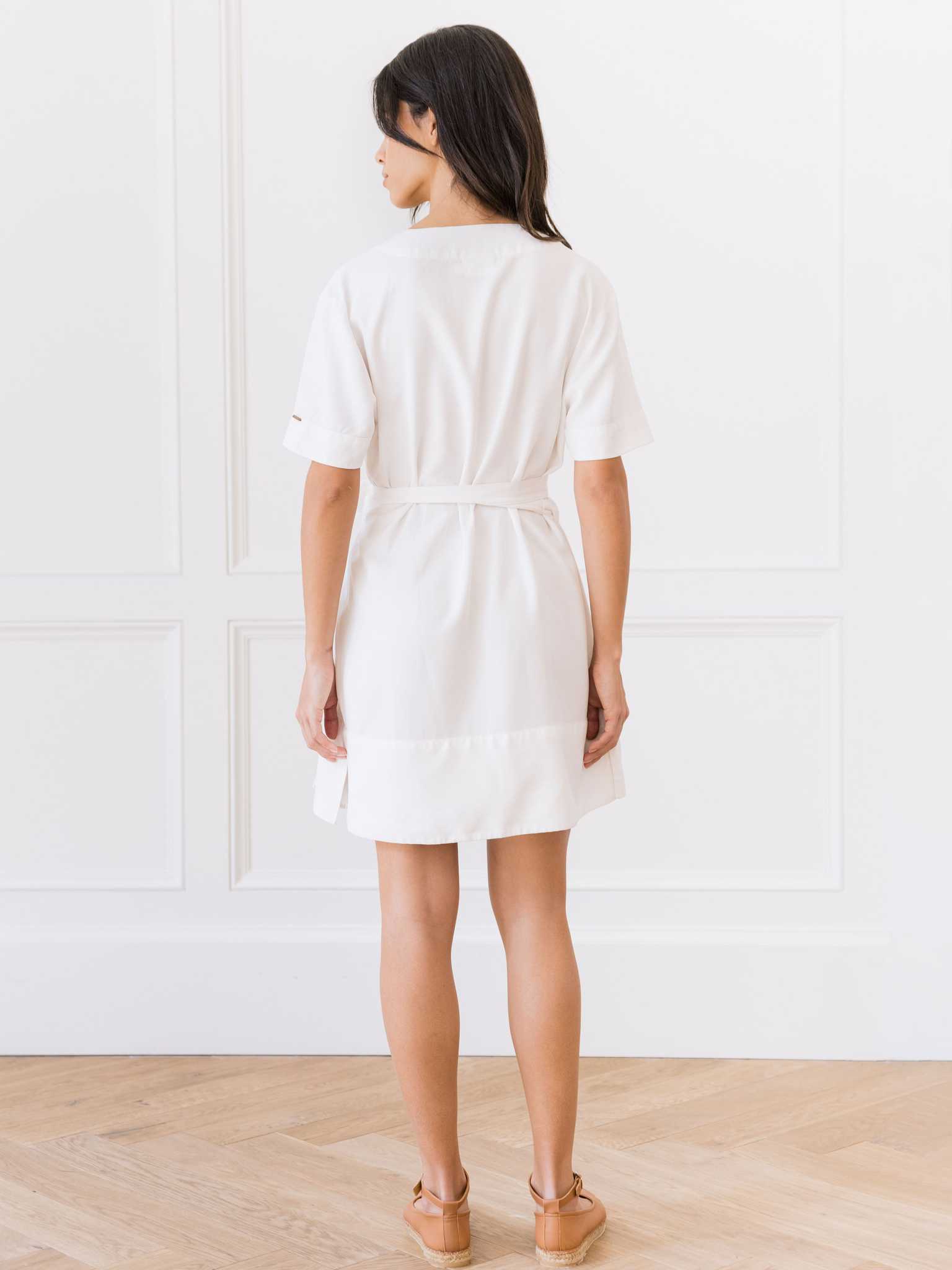 A person with long dark hair stands facing away, wearing the Women's Cozy Earth Coastal Comfort Wrap Dress from Cozy Earth with brown shoes. They are in a room with white paneled walls and a hardwood floor. |Color:White