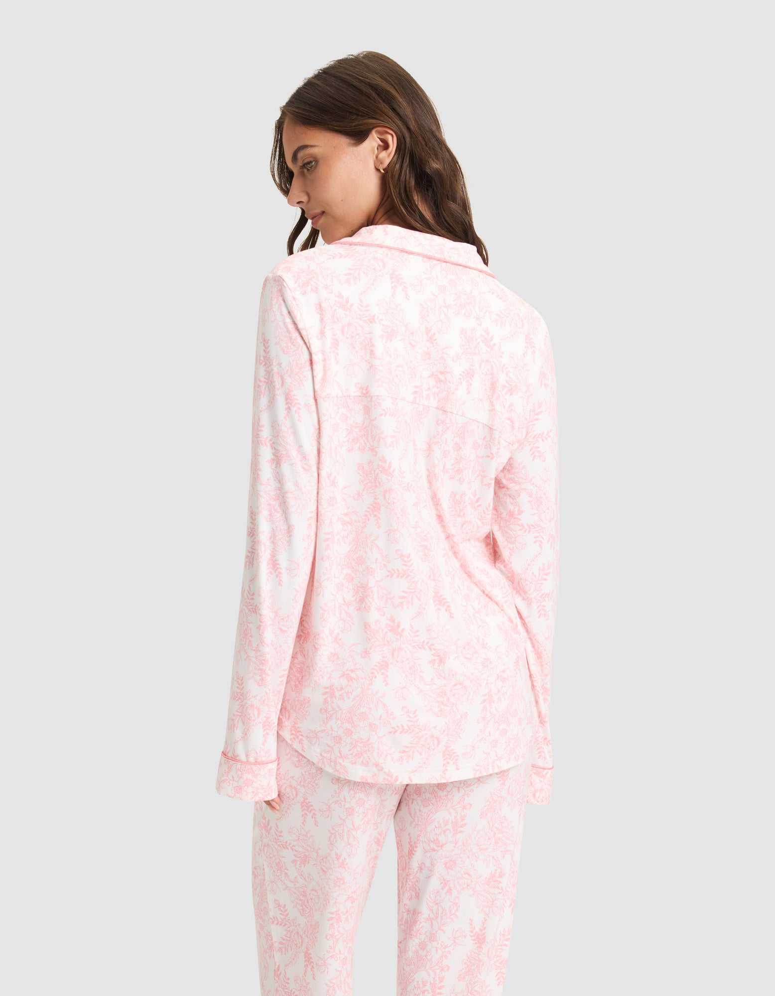 A woman models the Women's Bamboo Stretch-Knit Long Sleeve Pajama Set by Cozy Earth in light pink and white, featuring a collared, long-sleeve top and loose-fit pants. She is shown from the back against a plain light gray background. |Color:Tulip Toile