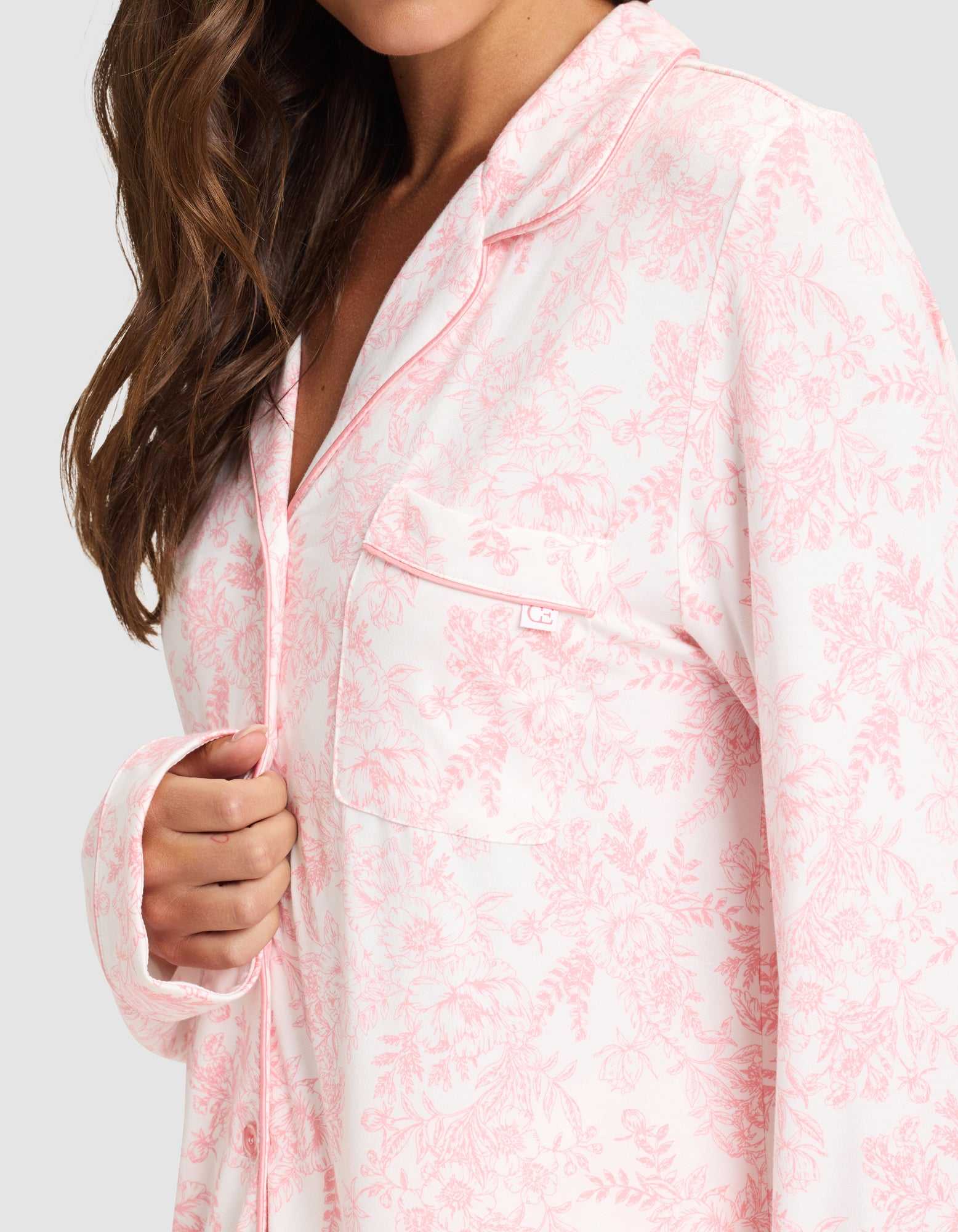 A woman with long brown hair wears the Cozy Earth Women's Bamboo Stretch-Knit Long Sleeve Pajama Set in a light pink and white floral pattern. She holds the top's edge, showing its chest pocket and piped trim. Her face is partially out of frame. |Color:Tulip Toile