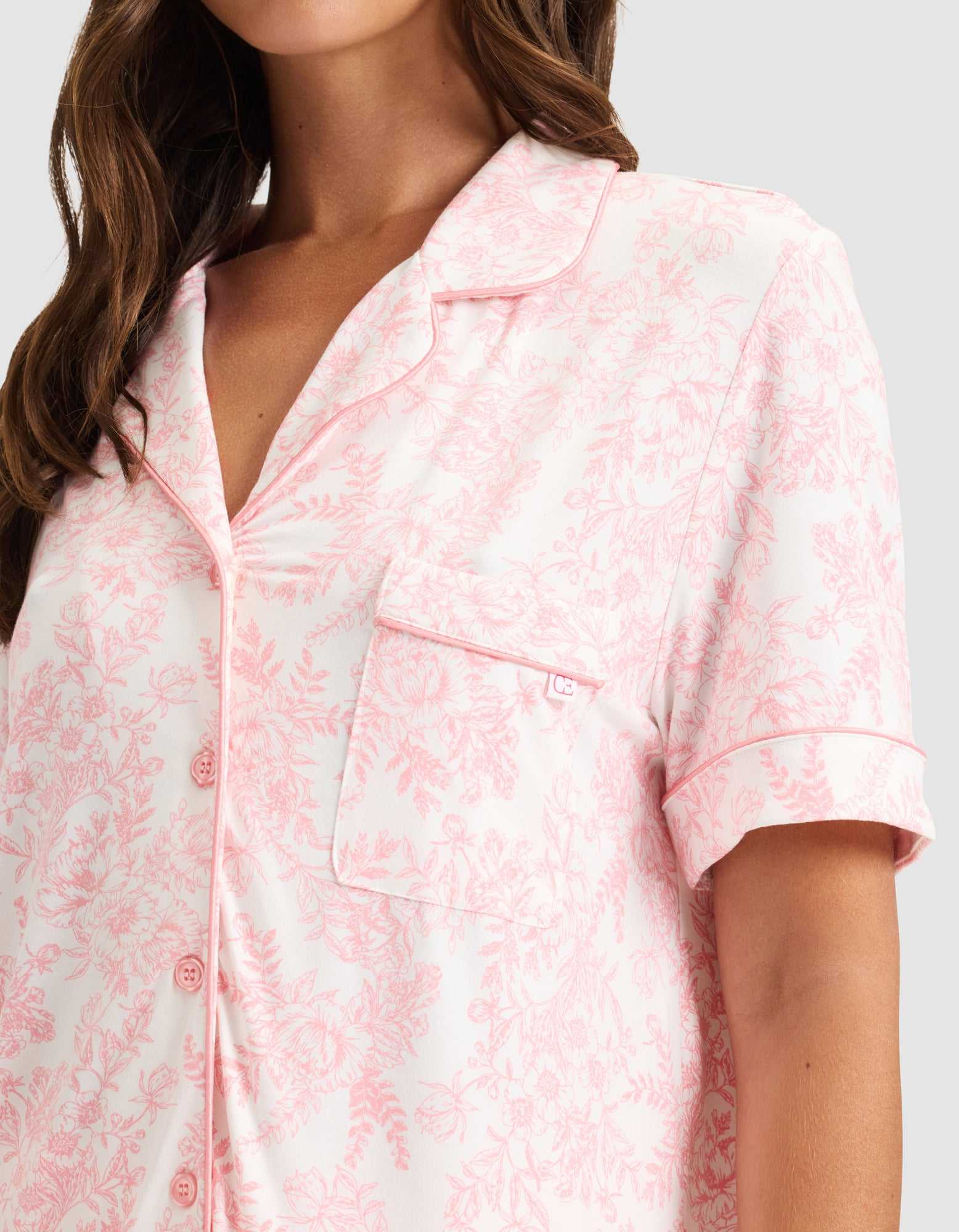 A woman wears HIDE's Women's Bamboo Stretch-Knit Short Sleeve Pajama Top in light pink with a floral pattern, notched collar, chest pocket, front buttons, and pink piping. Only her upper body is visible. |Color:Tulip Toile