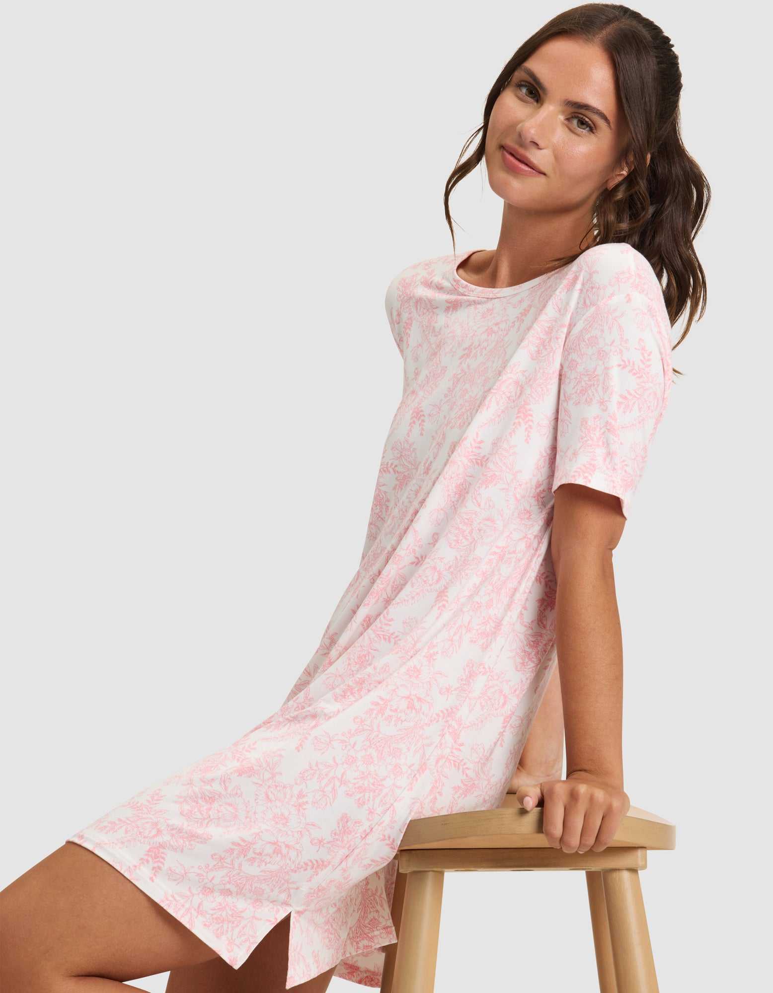 A woman with long brown hair sits sideways on a wooden stool, gently smiling at the camera while wearing Cozy Earth's Women's Bamboo Stretch-Knit Sleep Dress. |Color:Tulip Toile