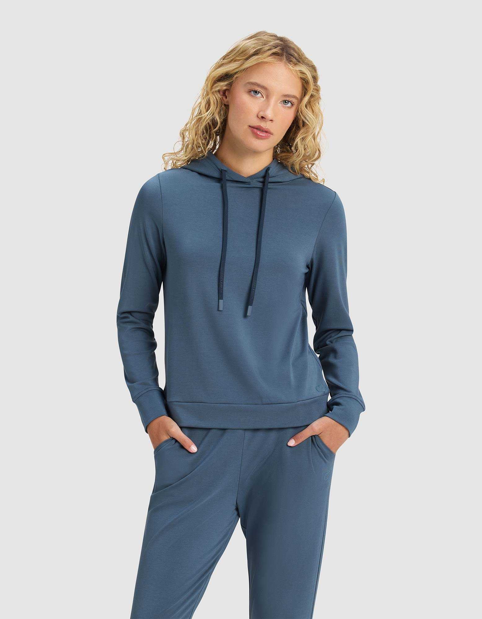 A woman with blonde curly hair models the Cozy Earth Women's Brushed Bamboo Hoodie in blue, paired with matching pants, standing hands-in-pockets against a plain light gray background.|Color:Dawn