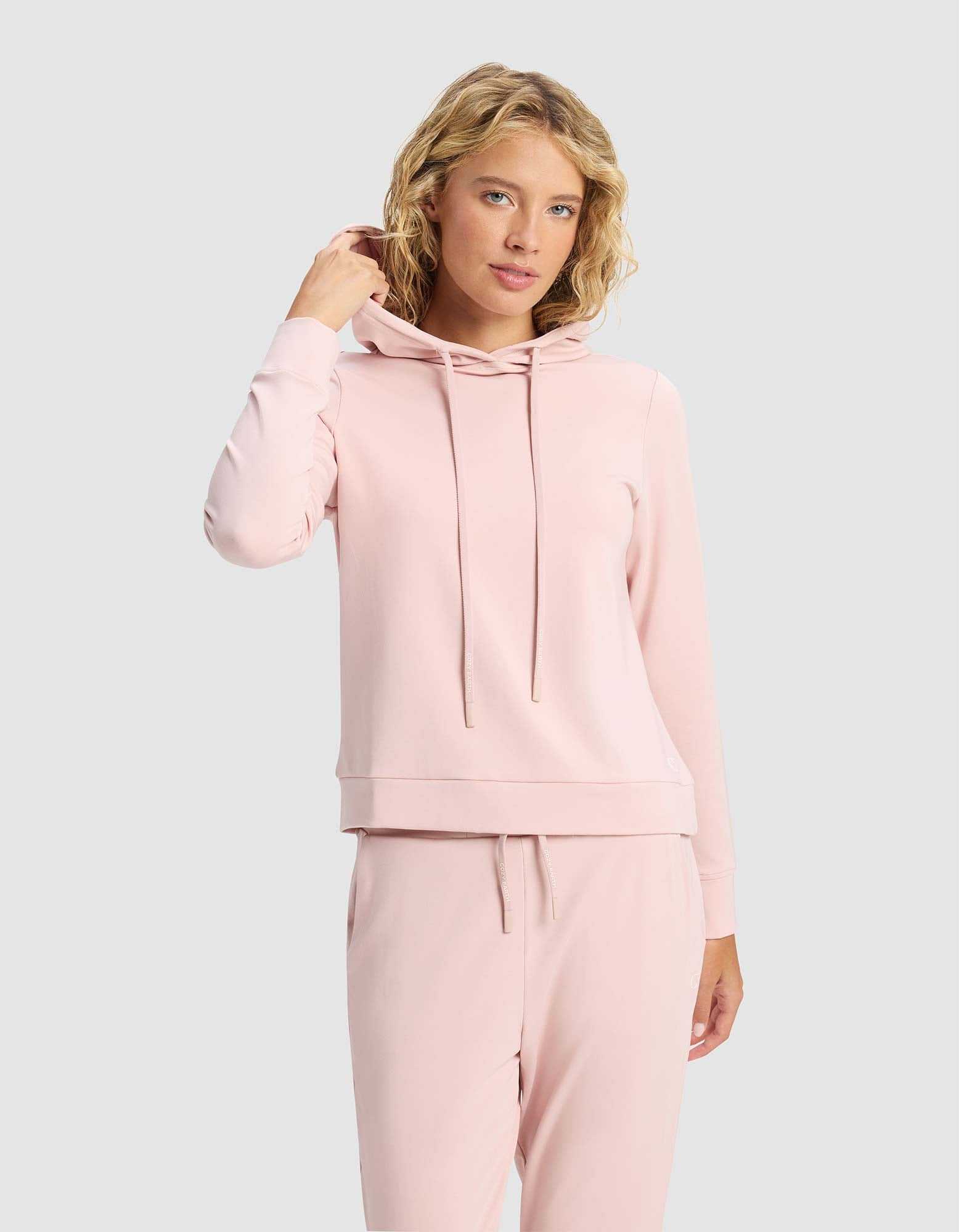 A woman with blond curly hair wears the Cozy Earth Women's Brushed Bamboo Hoodie in light pink, paired with matching sweatpants, against a plain light gray background. She faces the camera and touches her hair with one hand.|Color:Dusty Rose