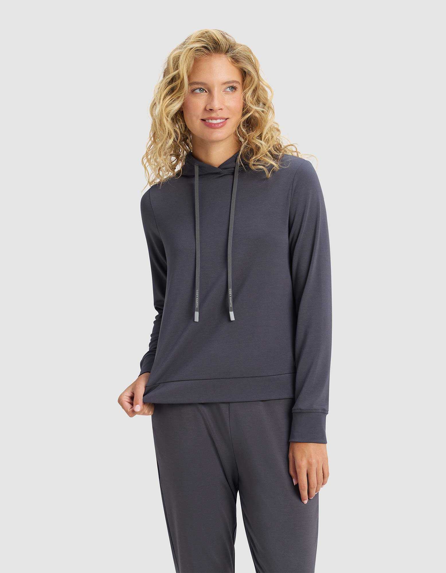 A woman with blonde, wavy hair stands against a light gray background, smiling softly while holding the hem of her Cozy Earth Women's Brushed Bamboo Hoodie, paired with matching pants.|Color:Graphite