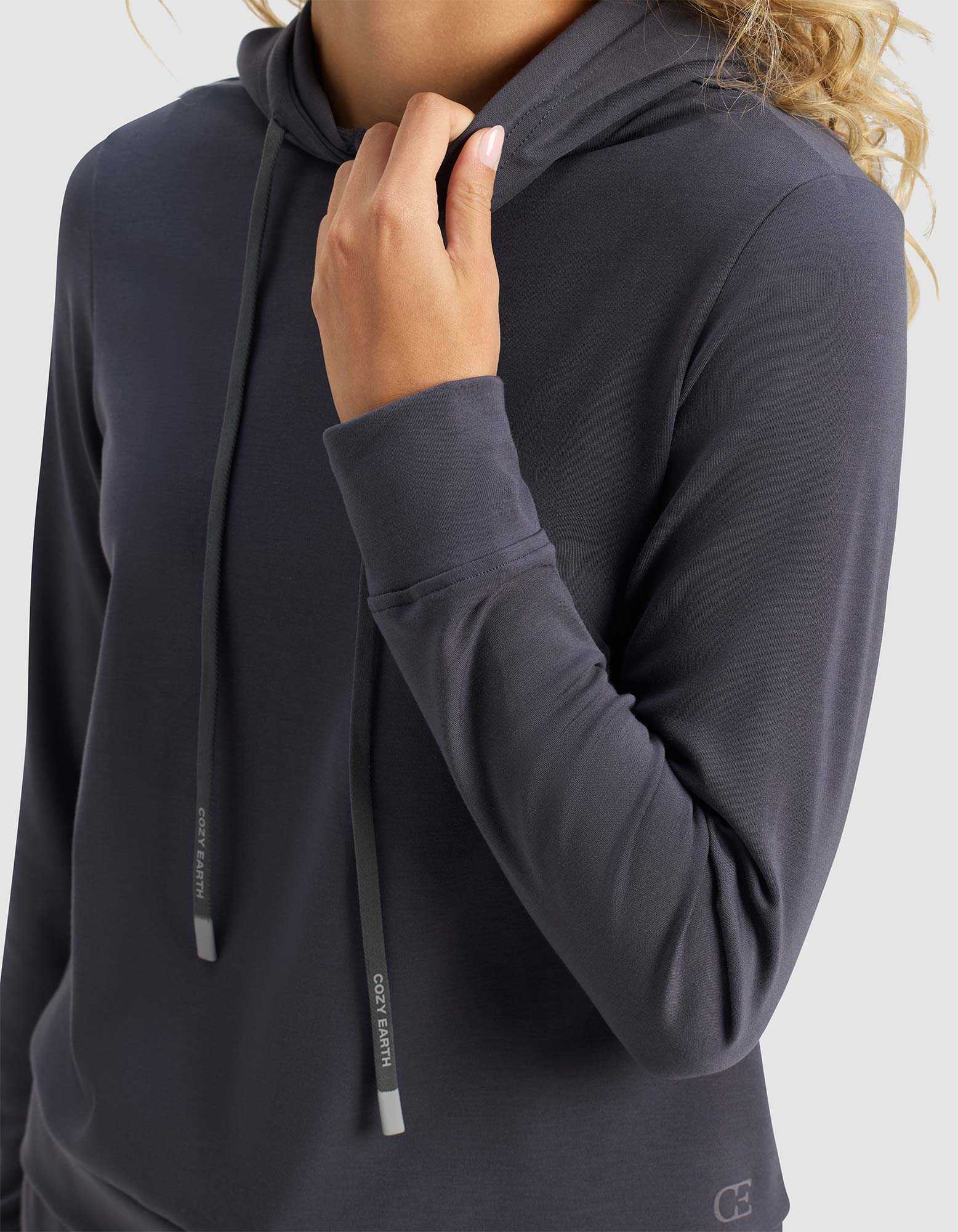 A person models the Cozy Earth Women's Brushed Bamboo Hoodie in dark gray, featuring long sleeves and a hood with drawstrings. They're holding the collar with one hand against a plain, light background; their face is not visible.|Color:Graphite