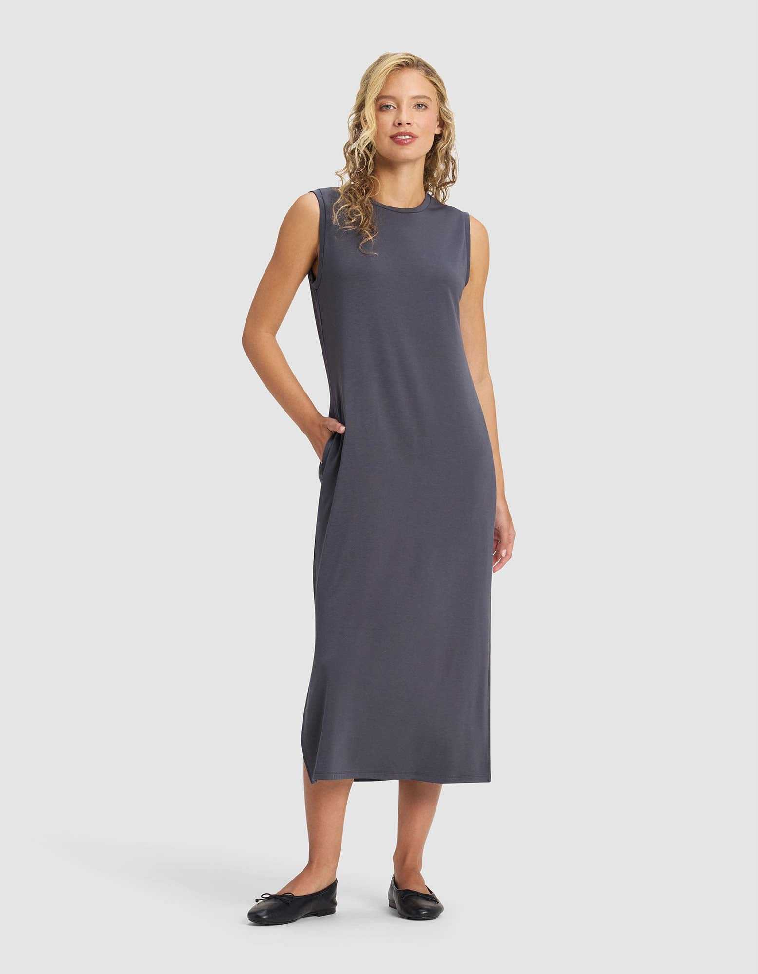 A woman with blonde curly hair wears the Cozy Earth Women's Brushed Bamboo Sleeveless Midi Dress in dark gray with black flats, standing against a plain light background, one hand in her pocket.|Color:Graphite