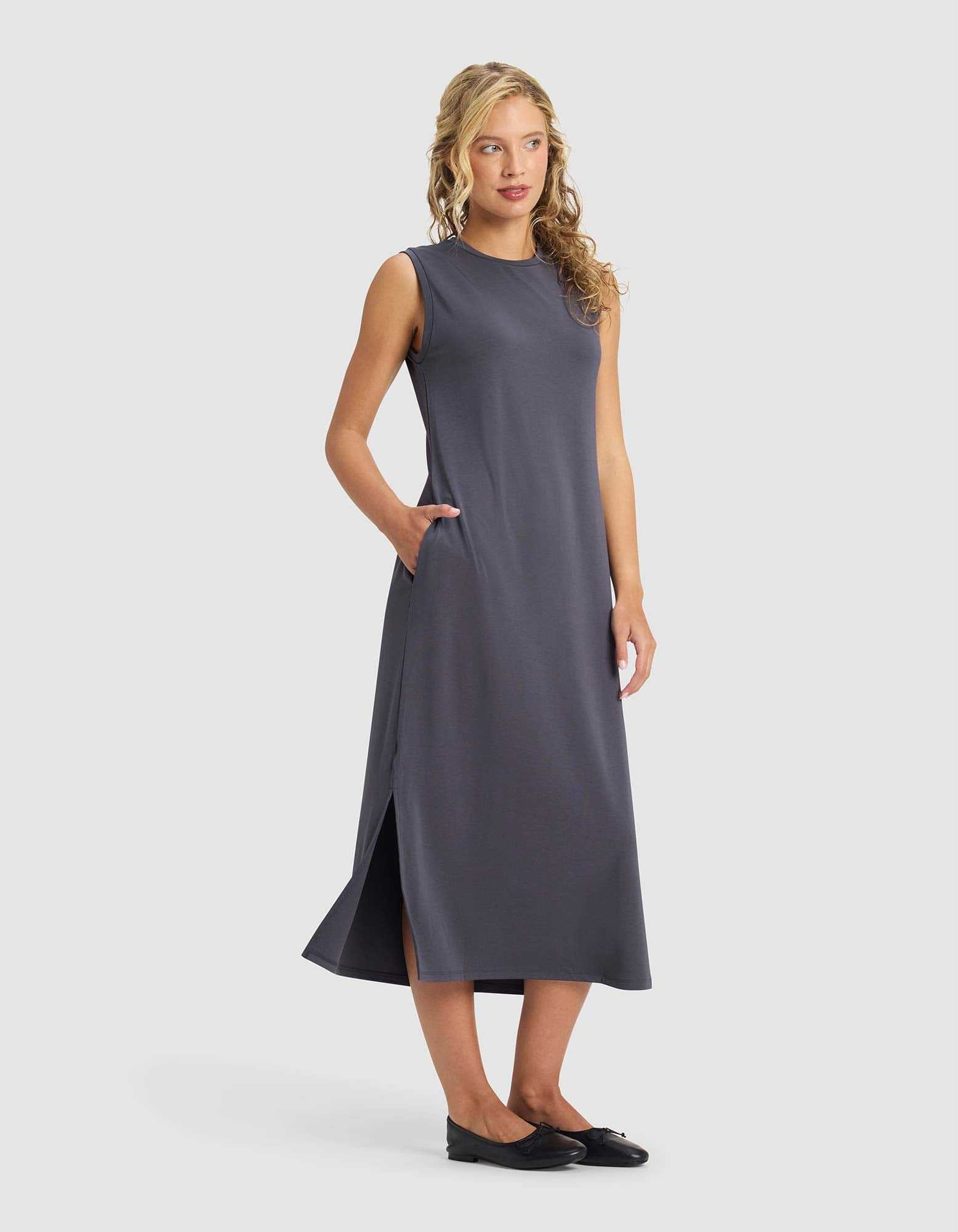 A woman with wavy blonde hair models the Cozy Earth Women's Brushed Bamboo Sleeveless Midi Dress in dark gray, featuring side pockets and a side slit, styled with black flats against a plain light gray background.|Color:Graphite