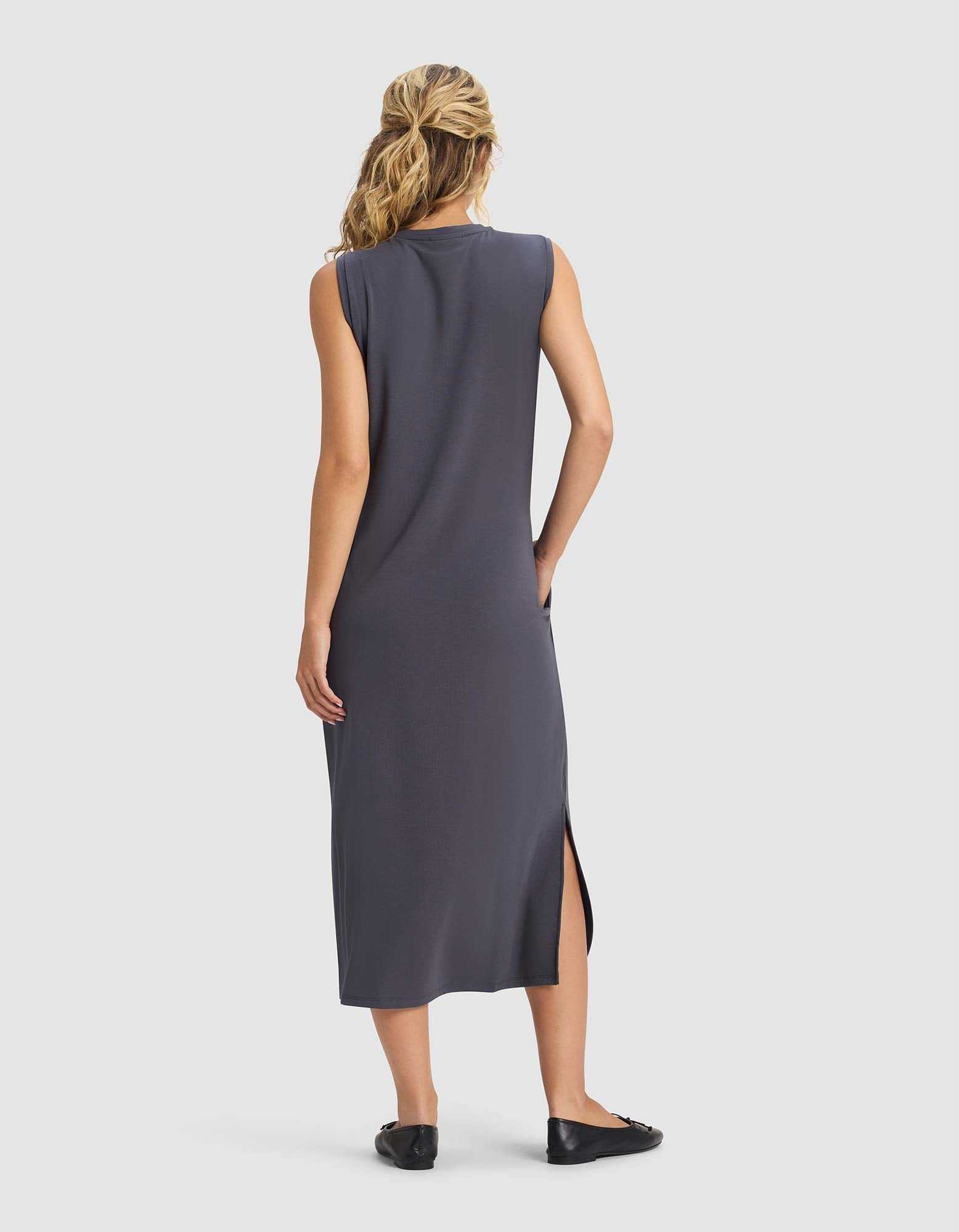 A woman with blonde hair in a ponytail faces away, wearing Cozy Earth's Women's Brushed Bamboo Sleeveless Midi Dress in dark gray with a side slit and black flats, against a plain white background.|Color:Graphite