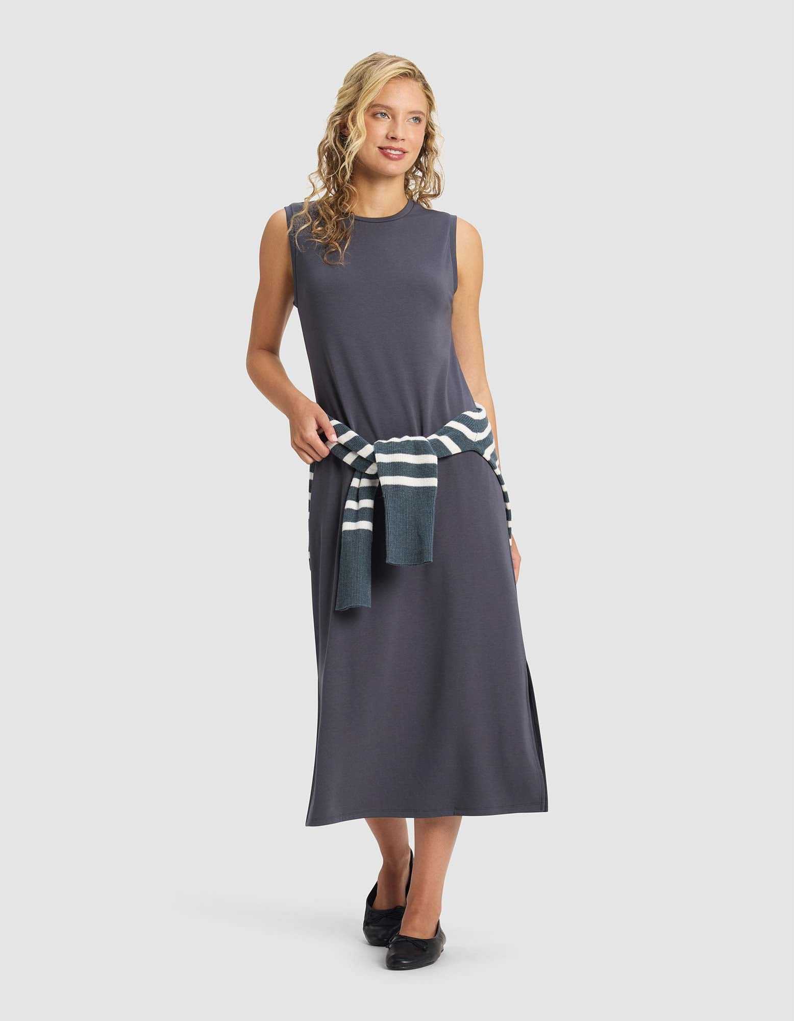 A woman with curly blonde hair smiles in front of a plain light background, wearing the Cozy Earth Women's Brushed Bamboo Sleeveless Midi Dress in dark gray with black shoes and a striped sweater tied at her waist.|Color:Graphite