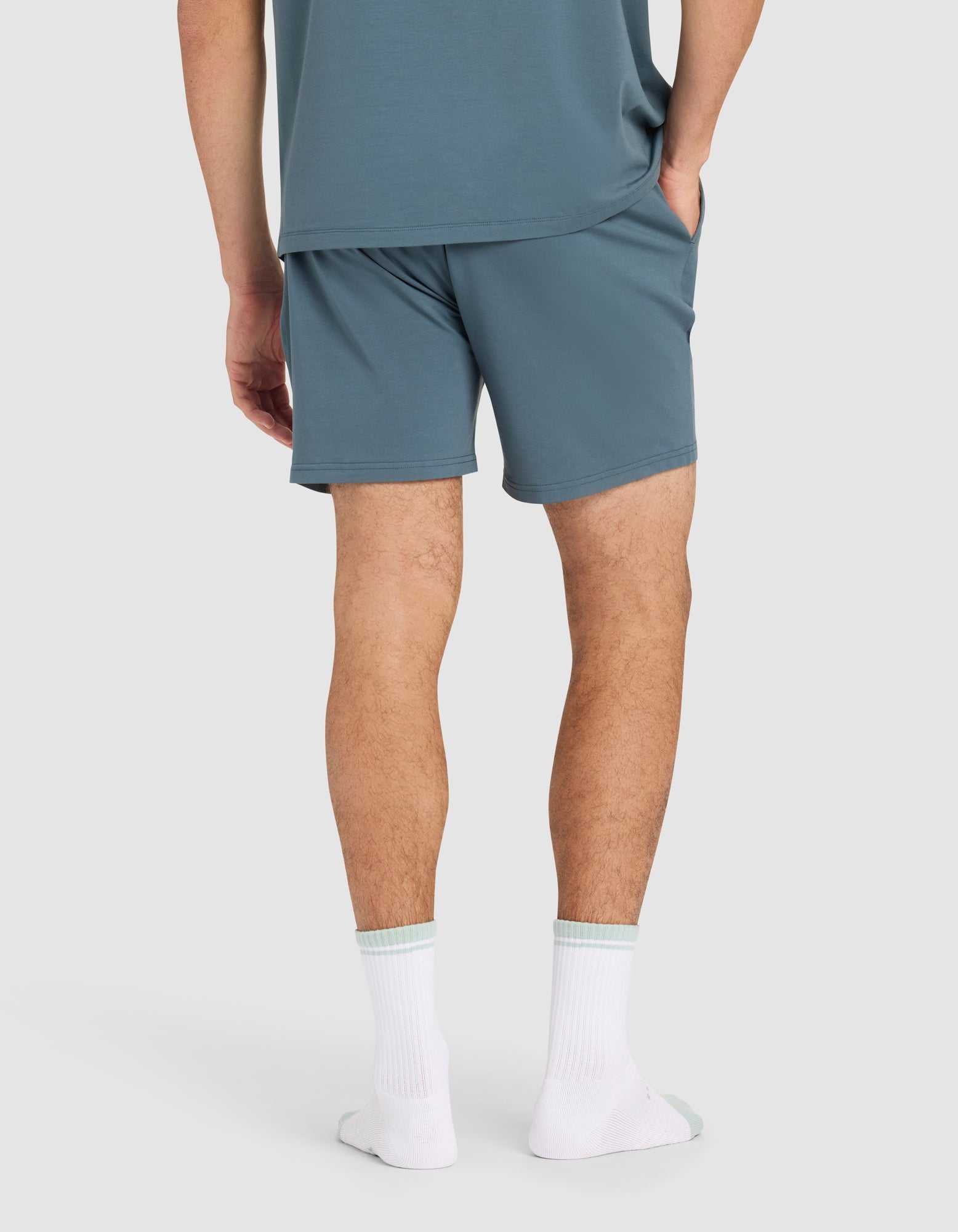 A person stands with hands in pockets, wearing Cozy Earth Men's First Layer Pajama Short in blue-green and a matching T-shirt, paired with white socks. The background is plain and light-colored.|Color:Dawn