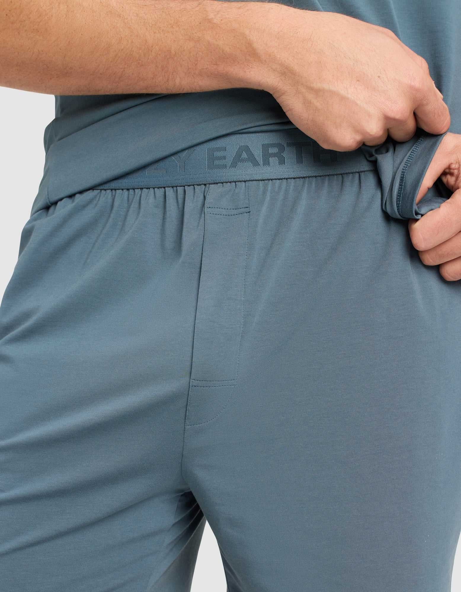 A person wearing Cozy Earth Men's First Layer Pajama Shorts slightly pulls the blue-gray waistband aside, revealing "EARTH" printed on the inner band. Only the lower torso and hand are visible.|Color:Dawn