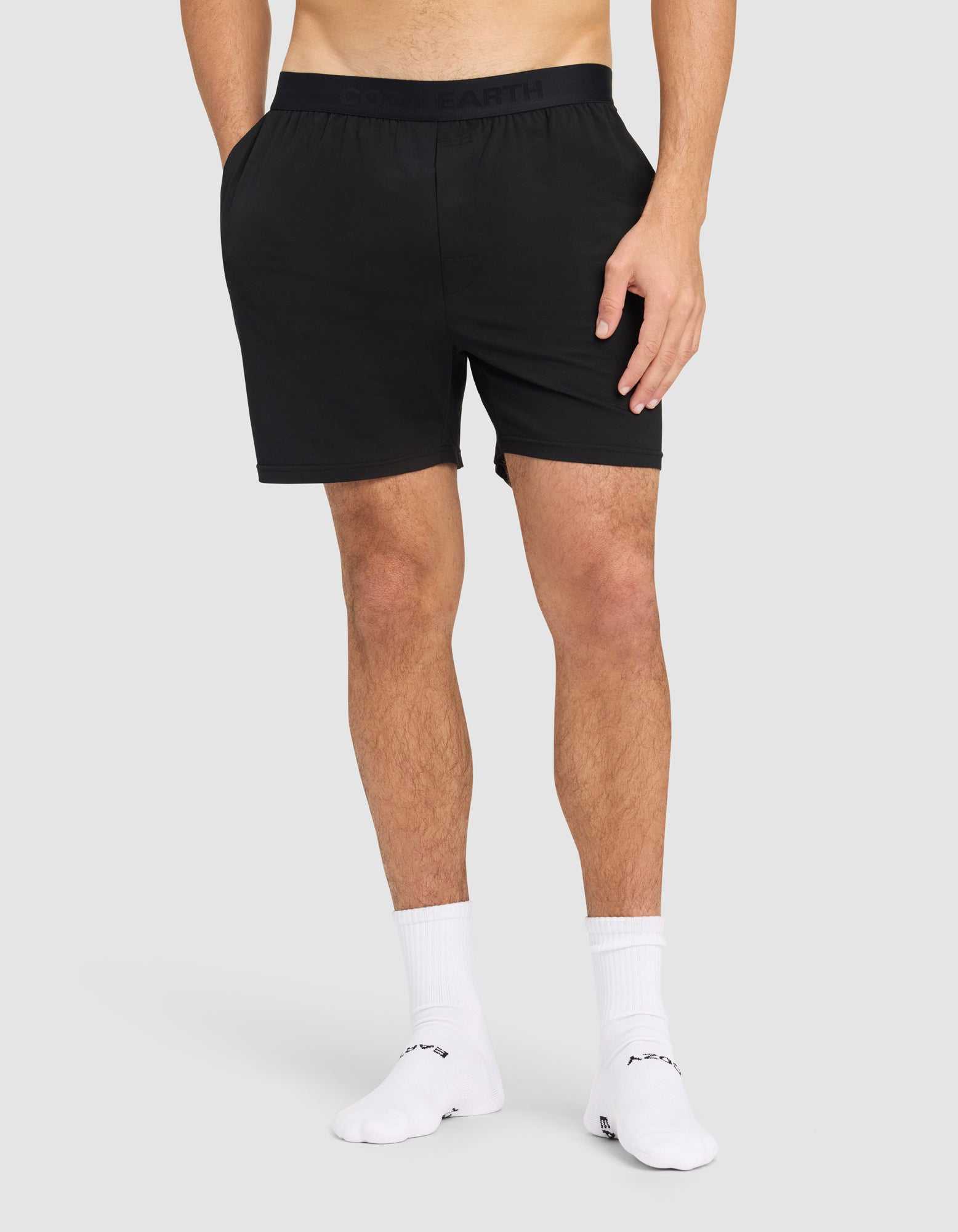 A man models Cozy Earth's Men's First Layer Pajama Short in black with white crew socks, standing on a plain light gray background. The image is cropped from mid-torso to feet, and his hands are relaxed at his sides. |Color:Jet Black