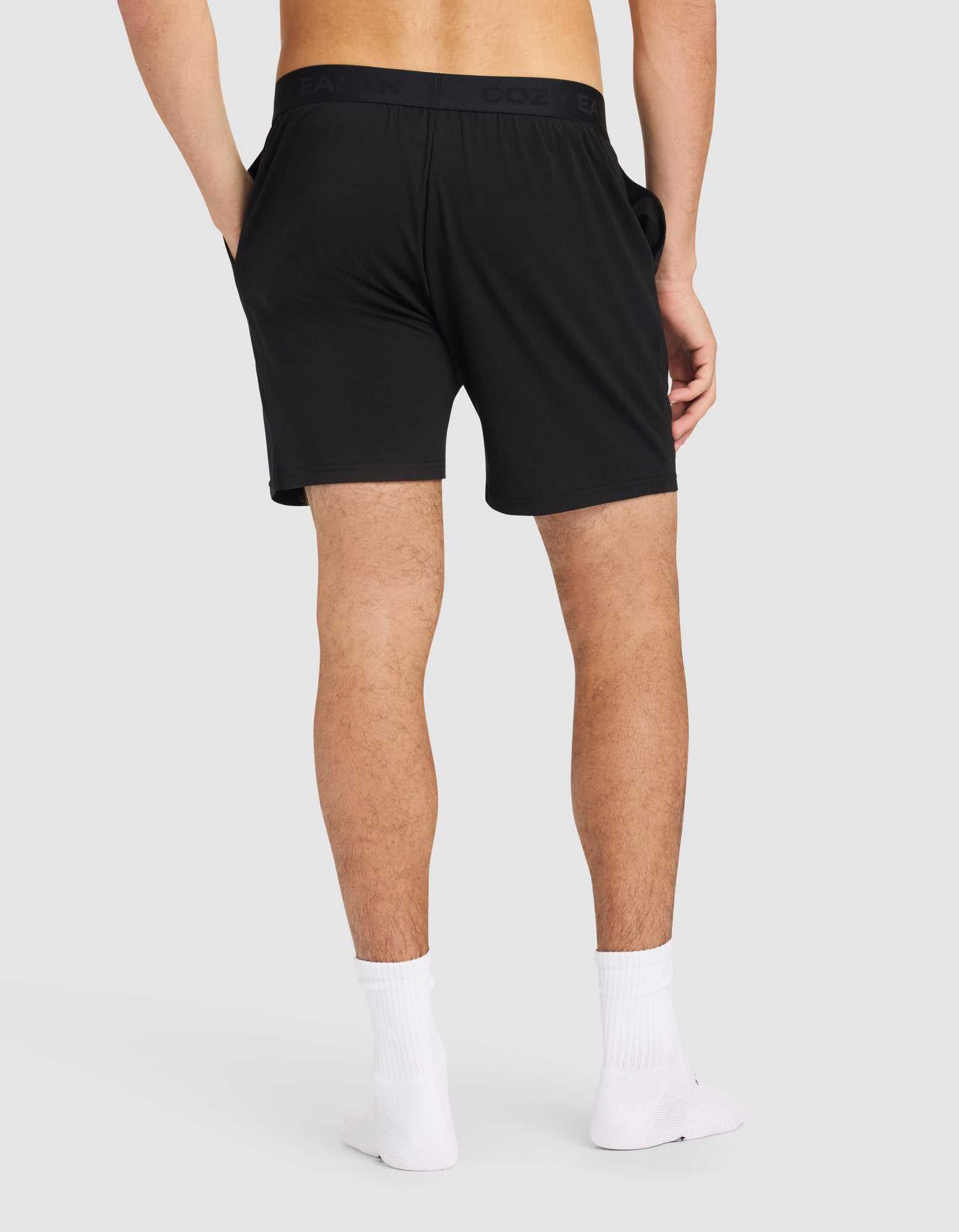 A man stands with his back to the camera, hands in the pockets of Cozy Earth Men's First Layer Pajama Shorts, paired with white ankle socks and no shoes, against a plain light gray background.|Color:Jet Black.