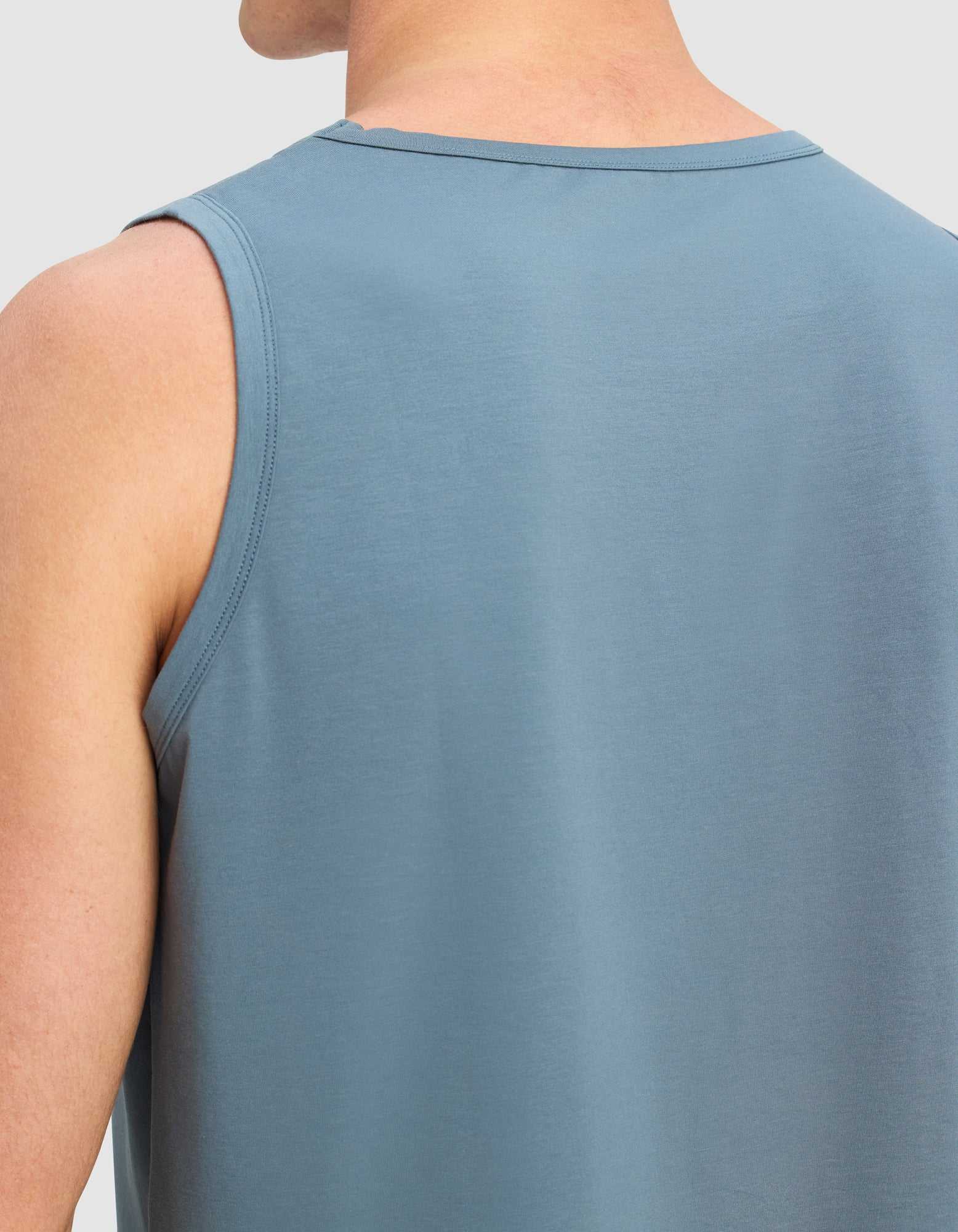 A close-up of a person wearing Cozy Earth's Men's First Layer Pajama Tank, sleeveless and blue, highlighting the upper back and shoulder against a plain light background.|Color:Dawn