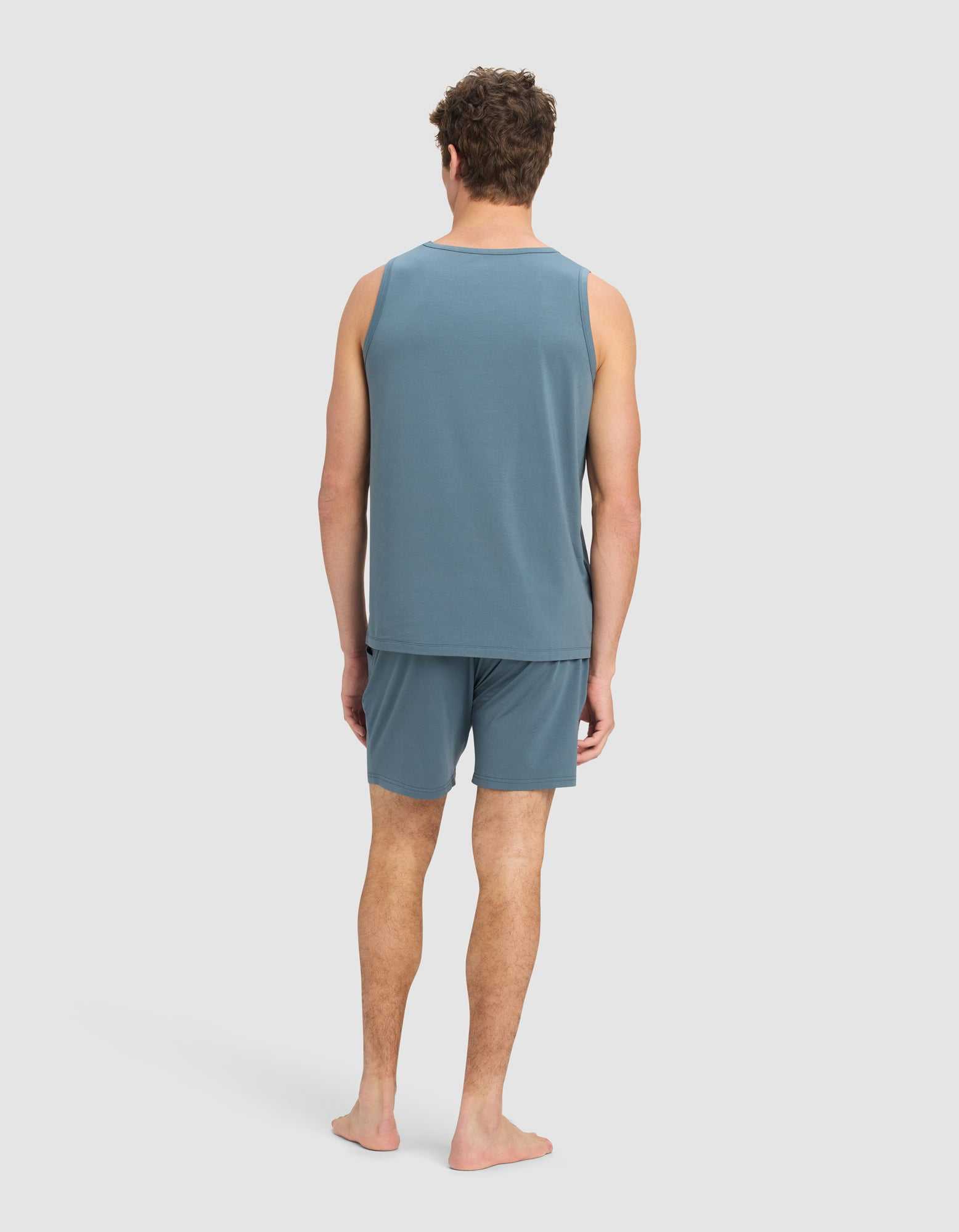 A man stands barefoot, facing away from the camera, wearing the Cozy Earth Men's First Layer Pajama Tank & Short Set in blue against a light gray background.