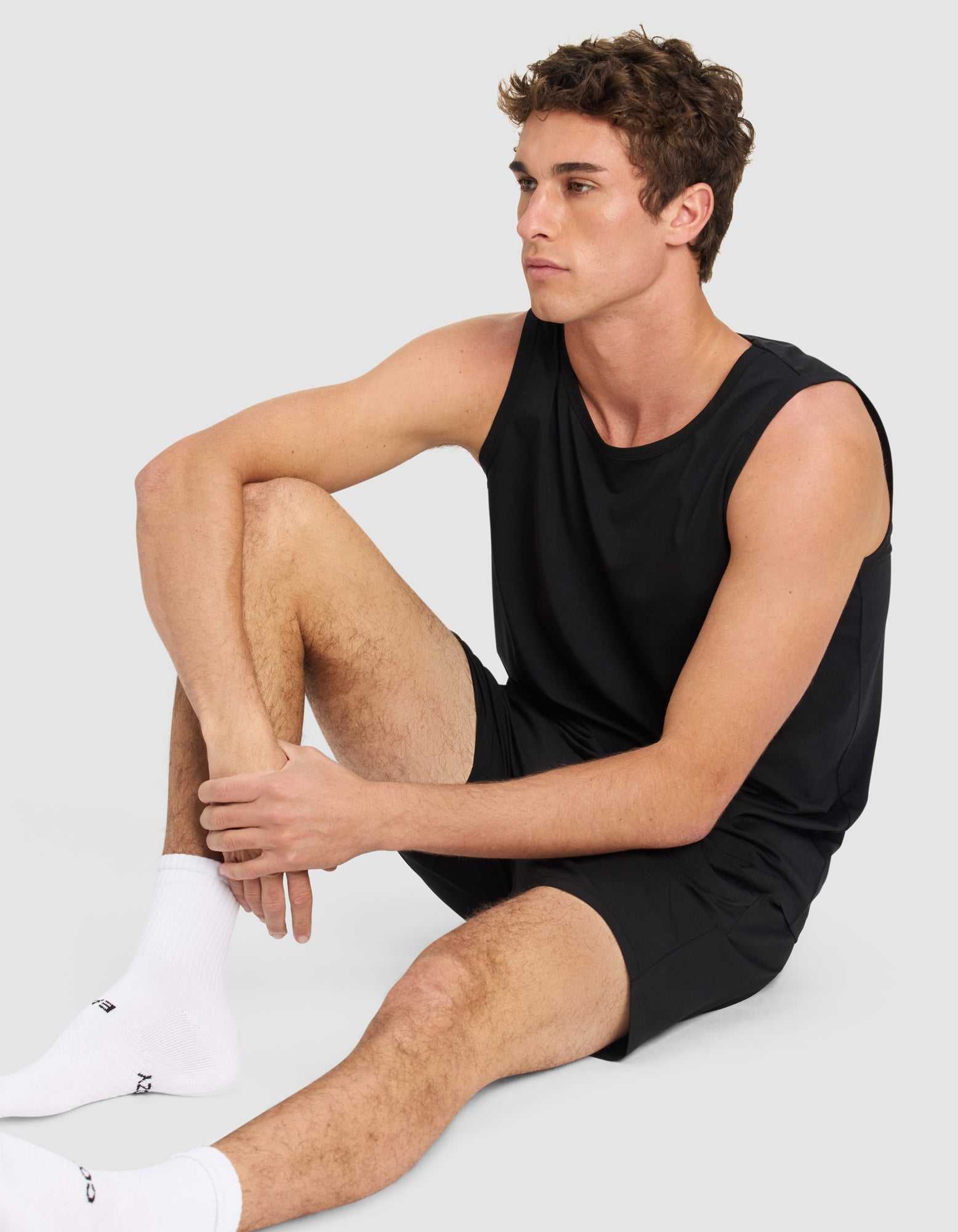 A young man with short brown hair sits on the floor against a light gray background, wearing the Men's First Layer Pajama Tank & Short Set by Cozy Earth. He looks to the side with a neutral expression, one knee raised.