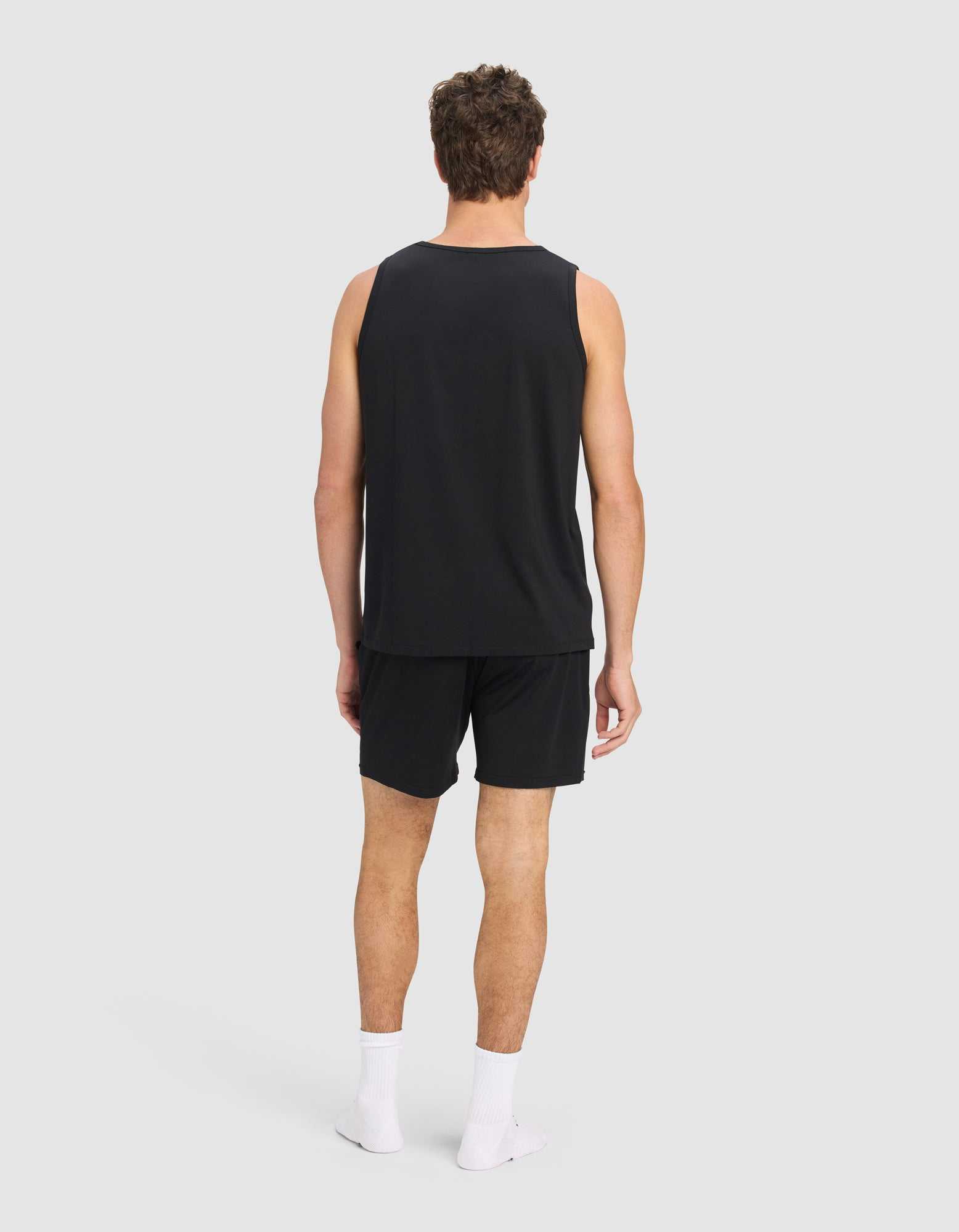 A man faces away from the camera, wearing Cozy Earth’s Men's First Layer Pajama Tank & Short Set in black, paired with white ankle socks, against a plain light gray background.