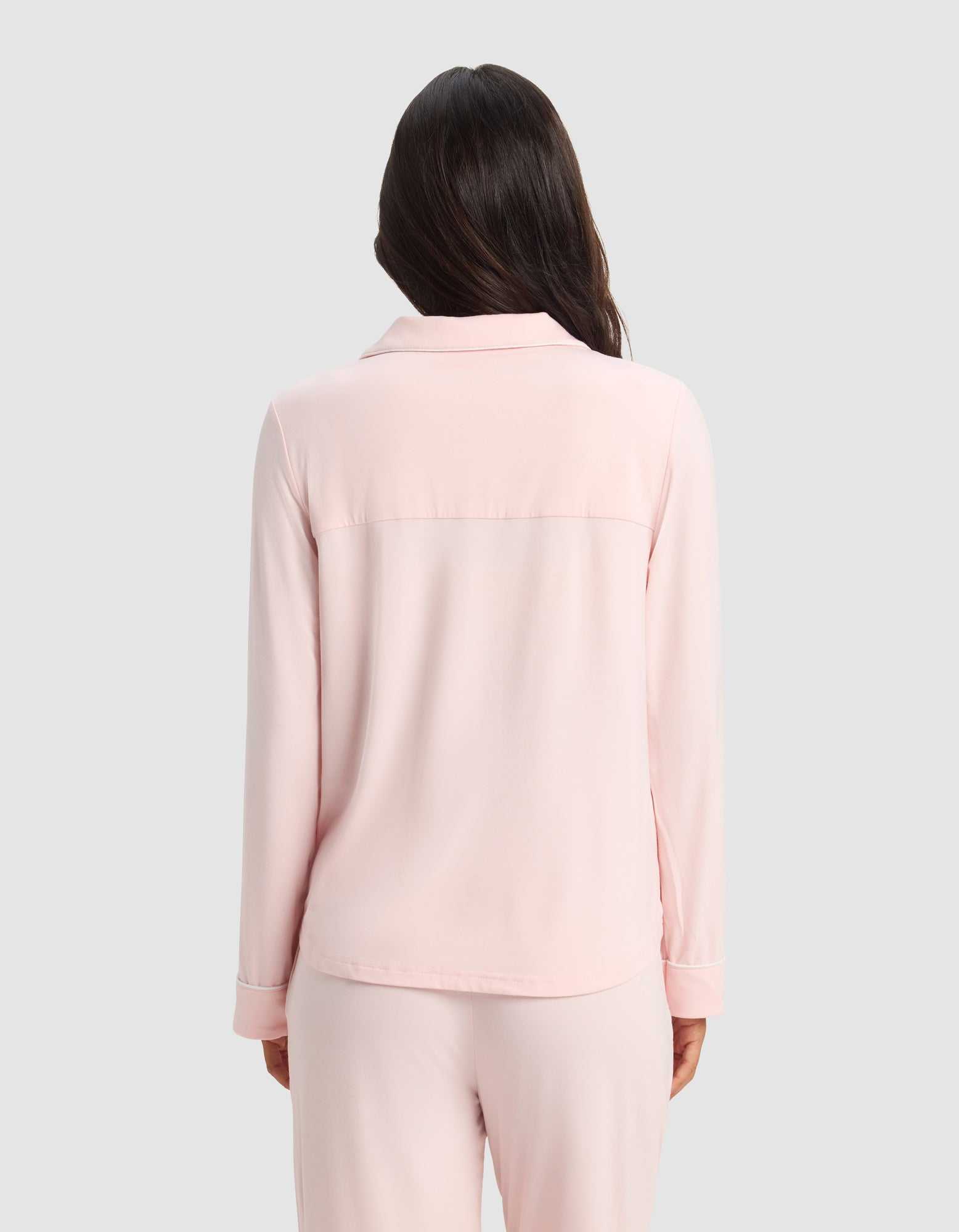 A woman with long dark hair stands with her back to the camera, wearing the Cozy Earth Women's Bamboo Stretch-Knit Long Sleeve Pajama Set in light pink against a plain light gray background.|Color:Blossom