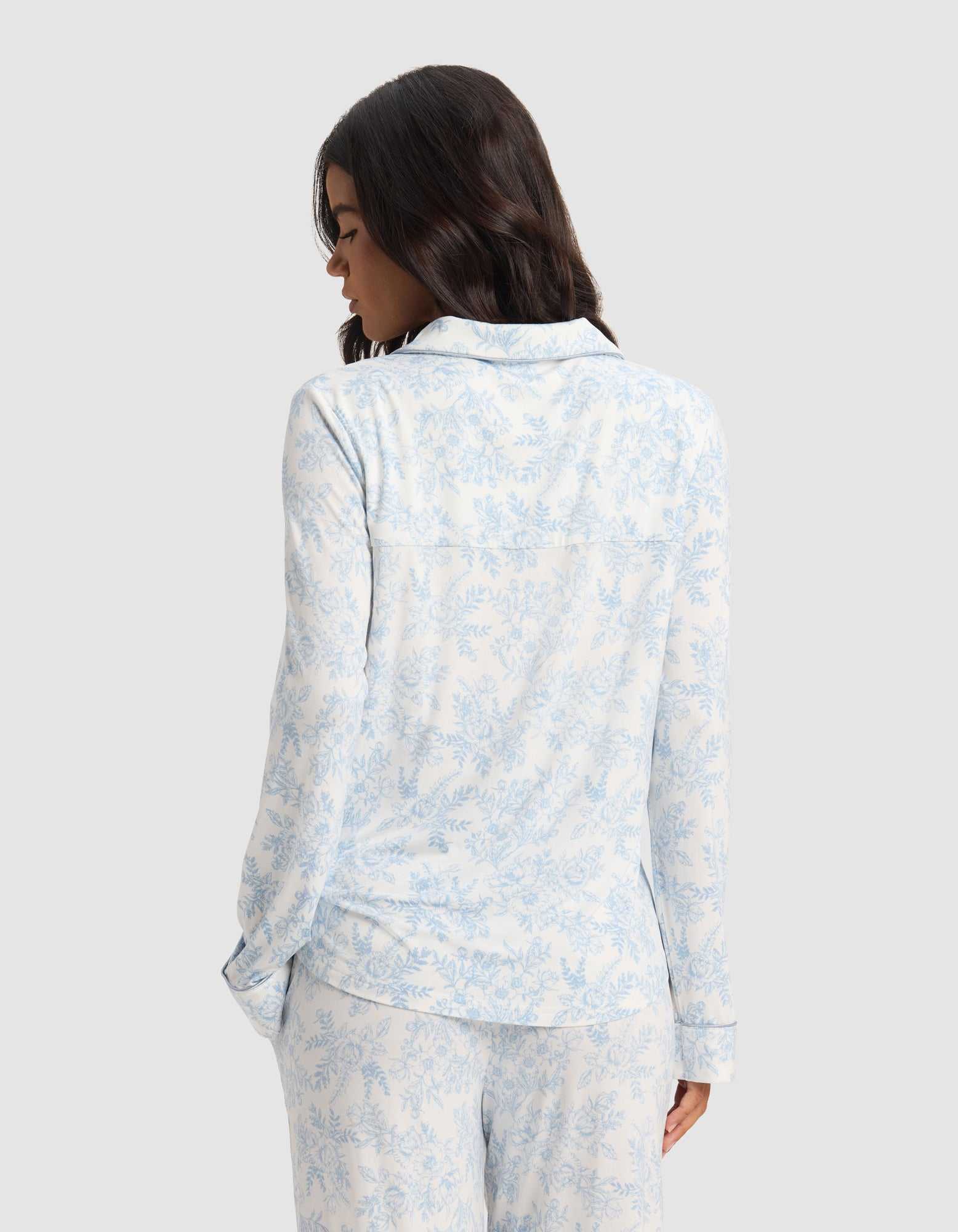 A woman with wavy dark hair is seen from behind, wearing the Cozy Earth Women's Bamboo Stretch-Knit Long Sleeve Pajama Set in a light blue and white floral pattern, standing against a plain light gray background.|Color:Sky Toile