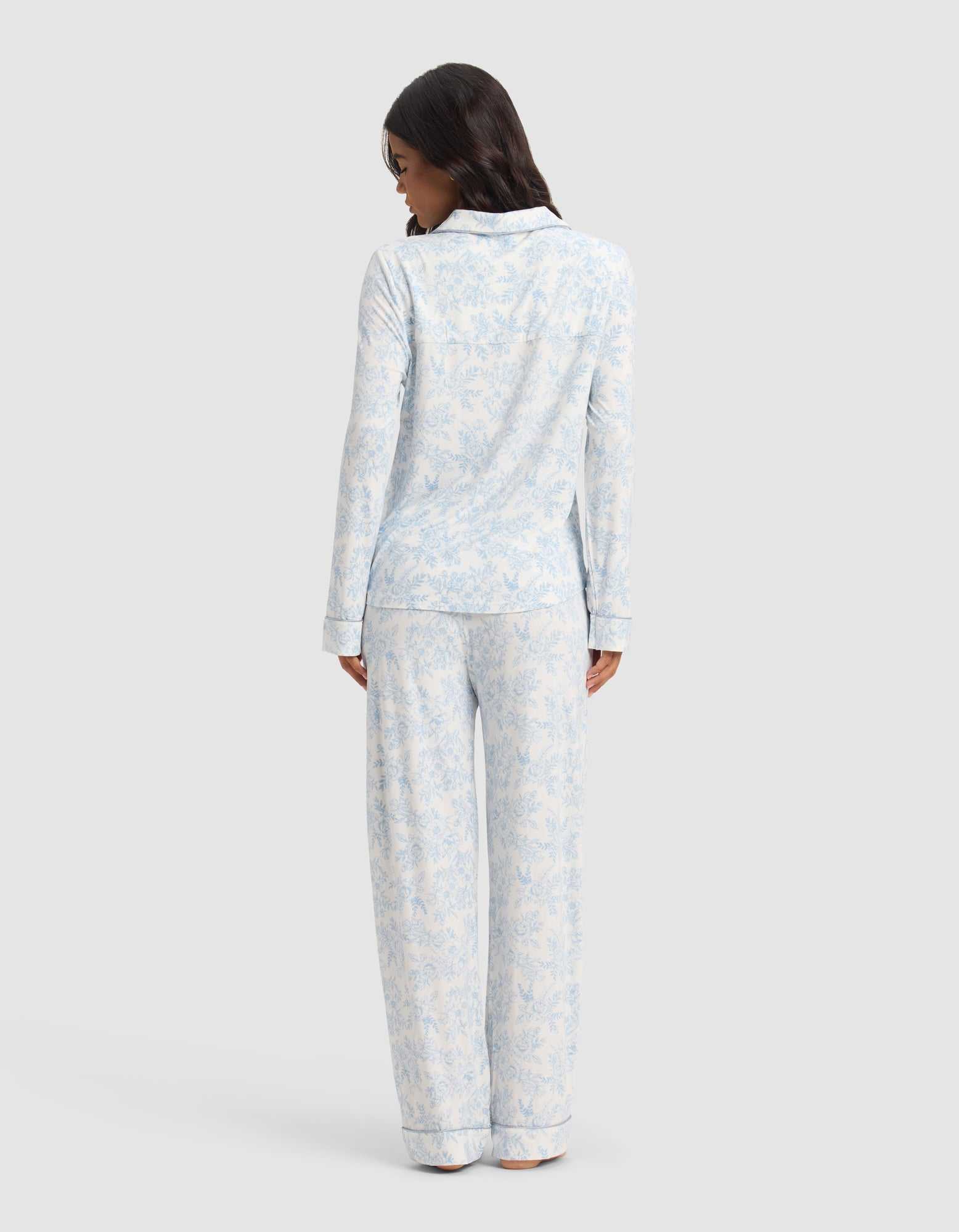 A woman faces away wearing the Cozy Earth Women's Bamboo Stretch-Knit Long Sleeve Pajama Set in light blue and white, featuring a delicate floral pattern, against a plain light background.|Color:Sky Toile